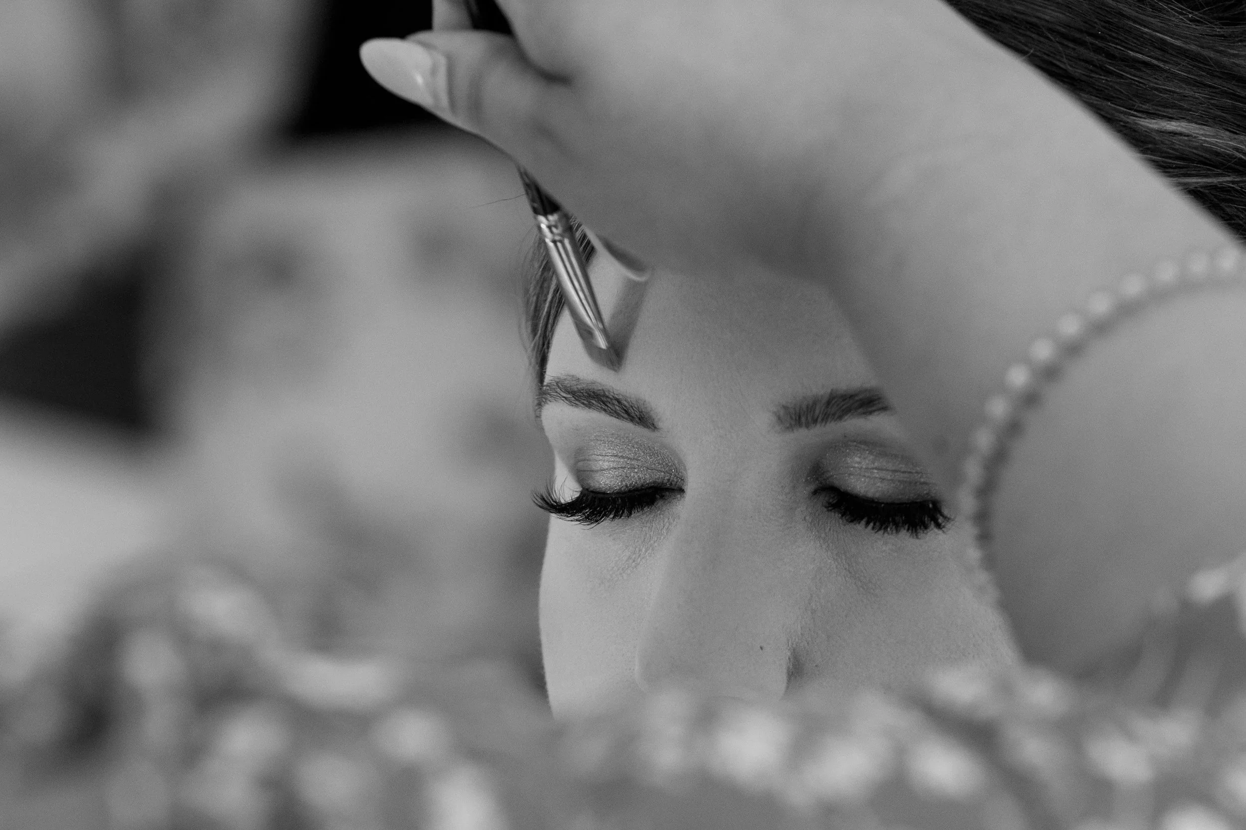 Close-up of a bride with eyes closed as an artist applies eye makeup with a brush.