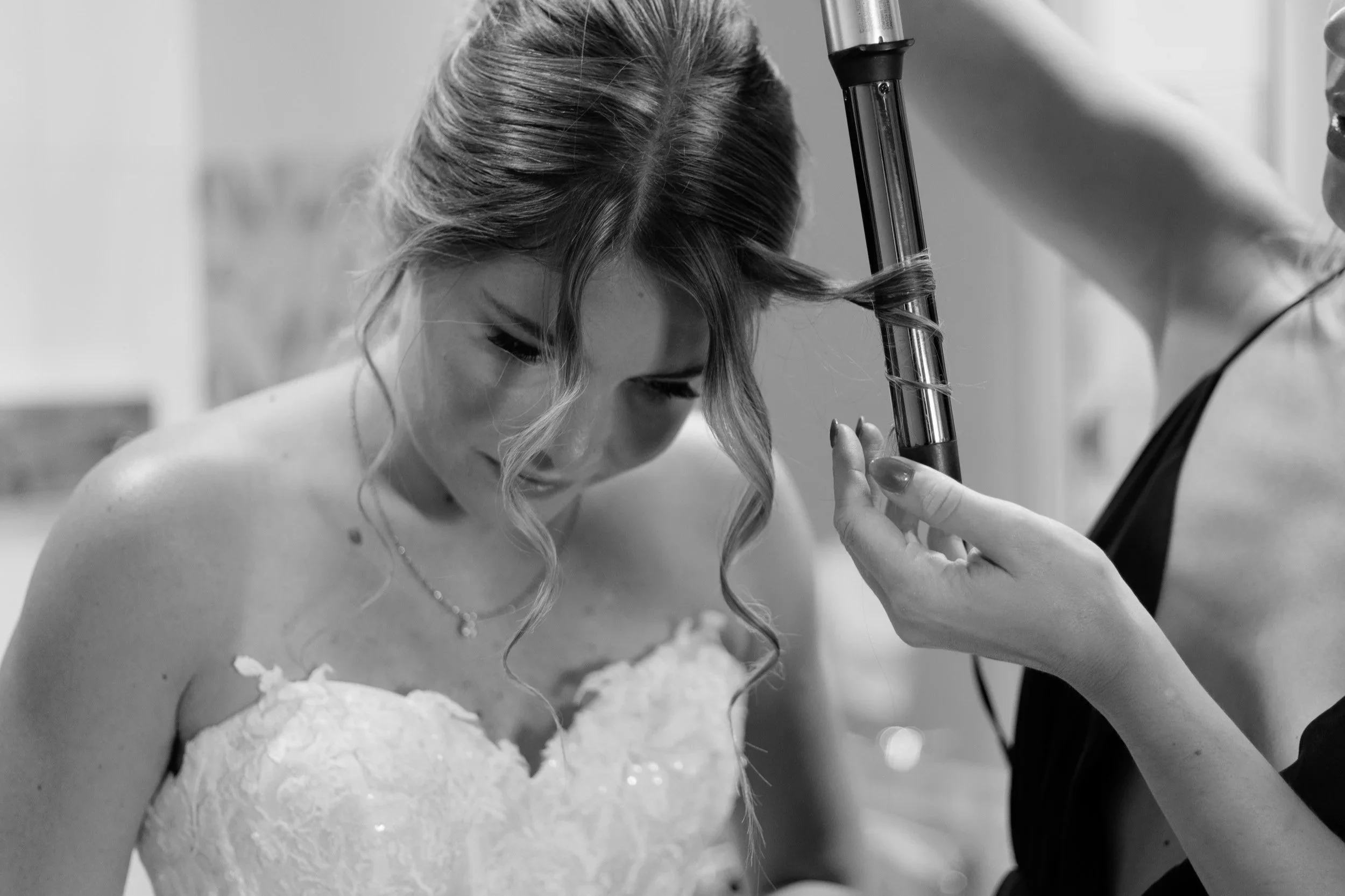 A bride with wavy hair in her wedding dress has her hair styled with hairspray by a stylist.