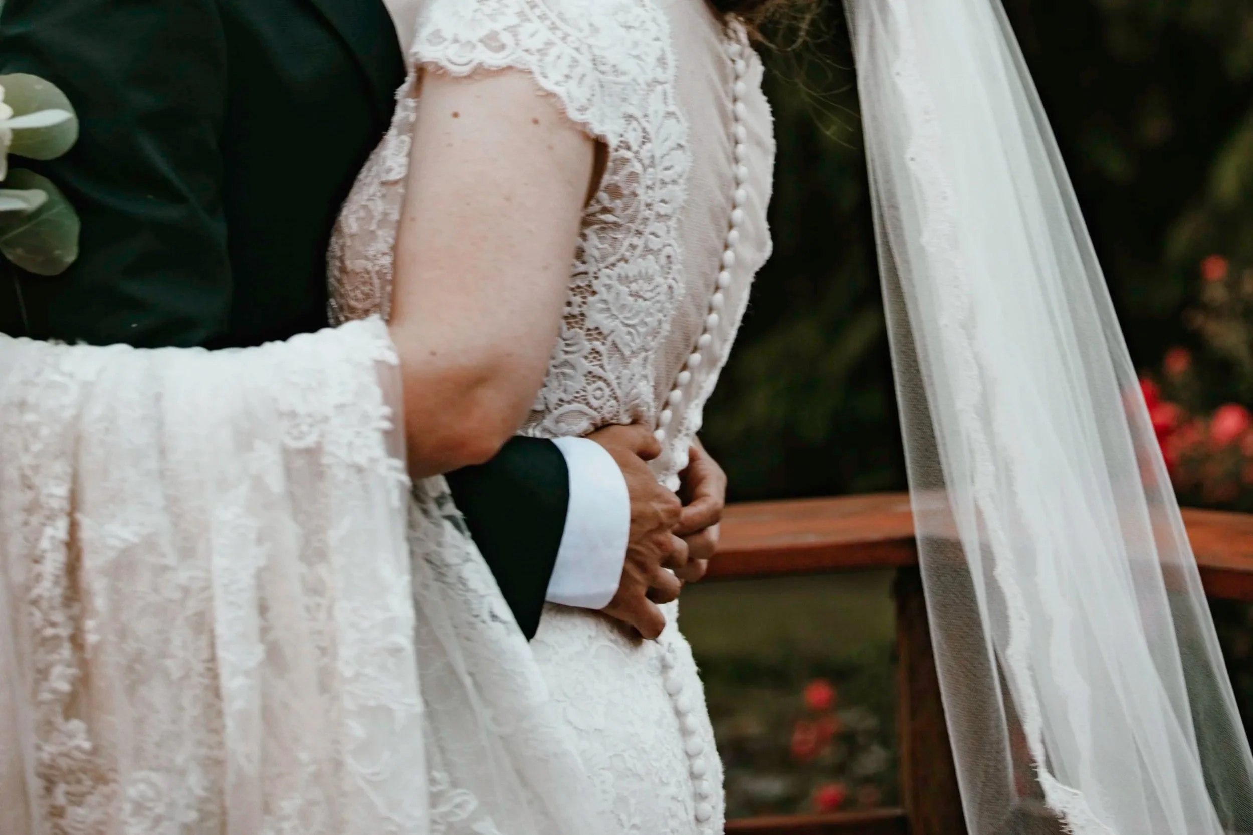 Close-up of a bride and groom holding hands during a wedding ceremony. The bride's dress has lace details and a veil. The groom is wearing a black suit with white shirt cuffs.
