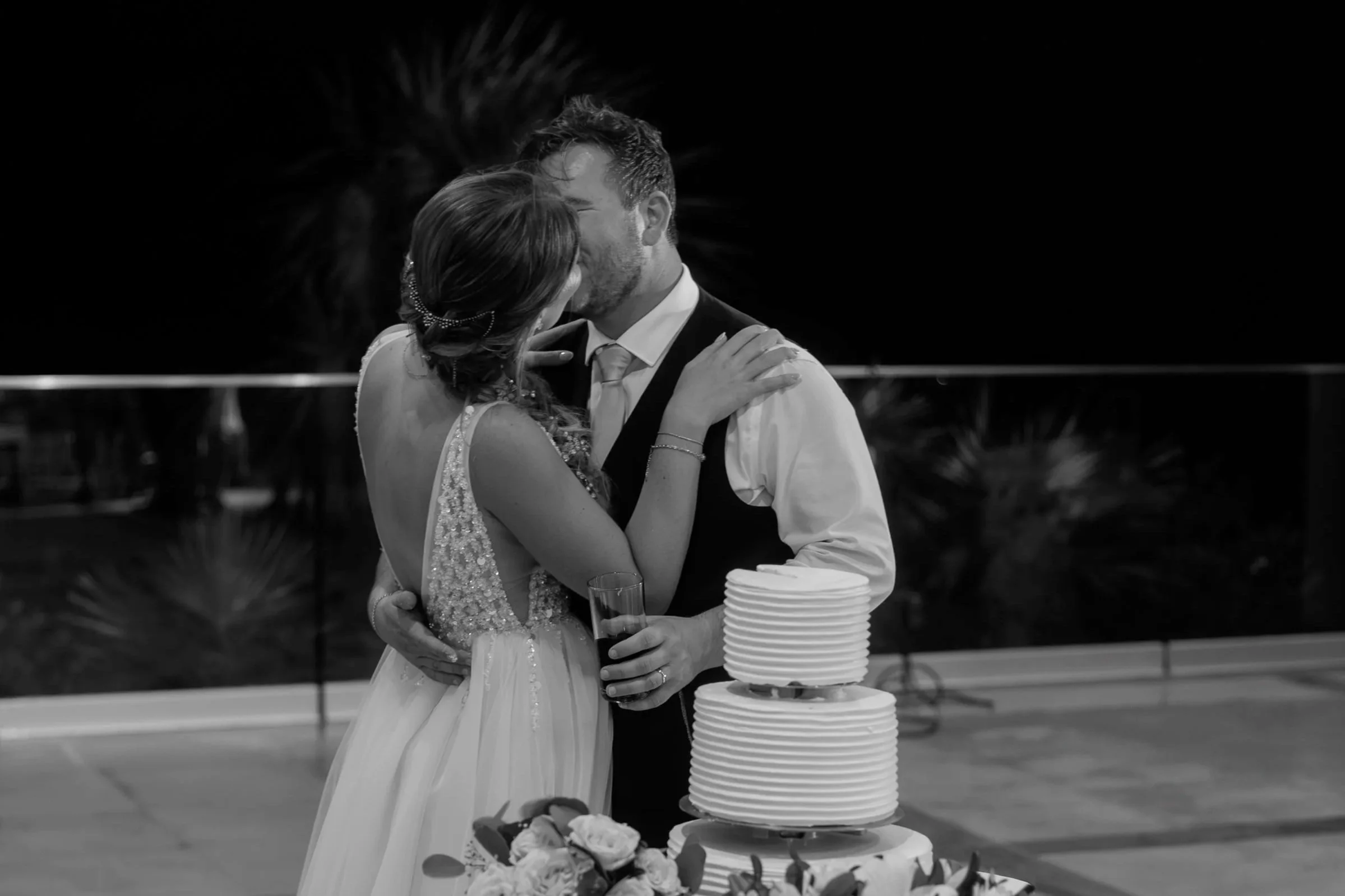 A black and white photo of a couple sharing a kiss at their wedding, with a wedding cake and flower decorations in front of them.