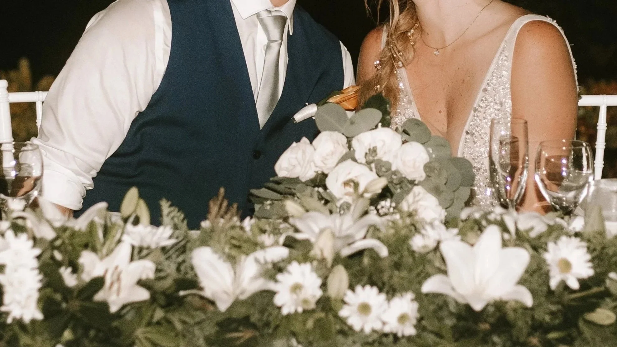 A bride and groom sitting at a wedding reception table decorated with white flowers, including lilies and daisies, surrounded by wine glasses and tableware. The bride is dressed in a sparkly gown, and the groom is in a dark vest with a white shirt an