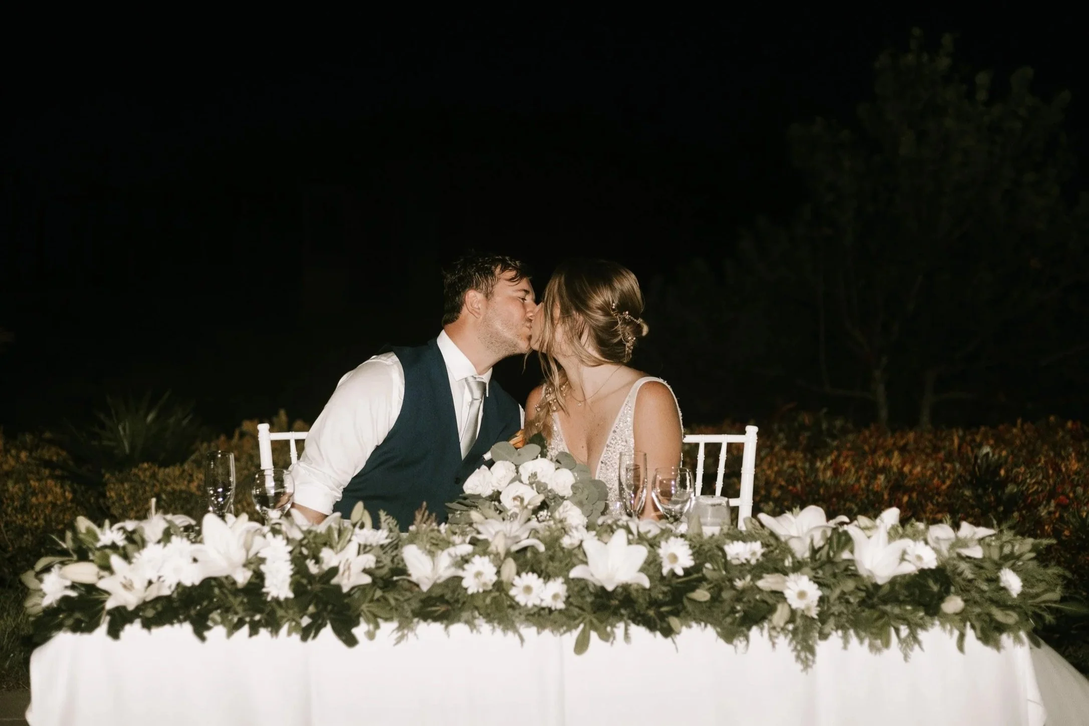 A bride and groom share a kiss at their wedding reception, sitting at a decorated table with white flowers and candles at night.
