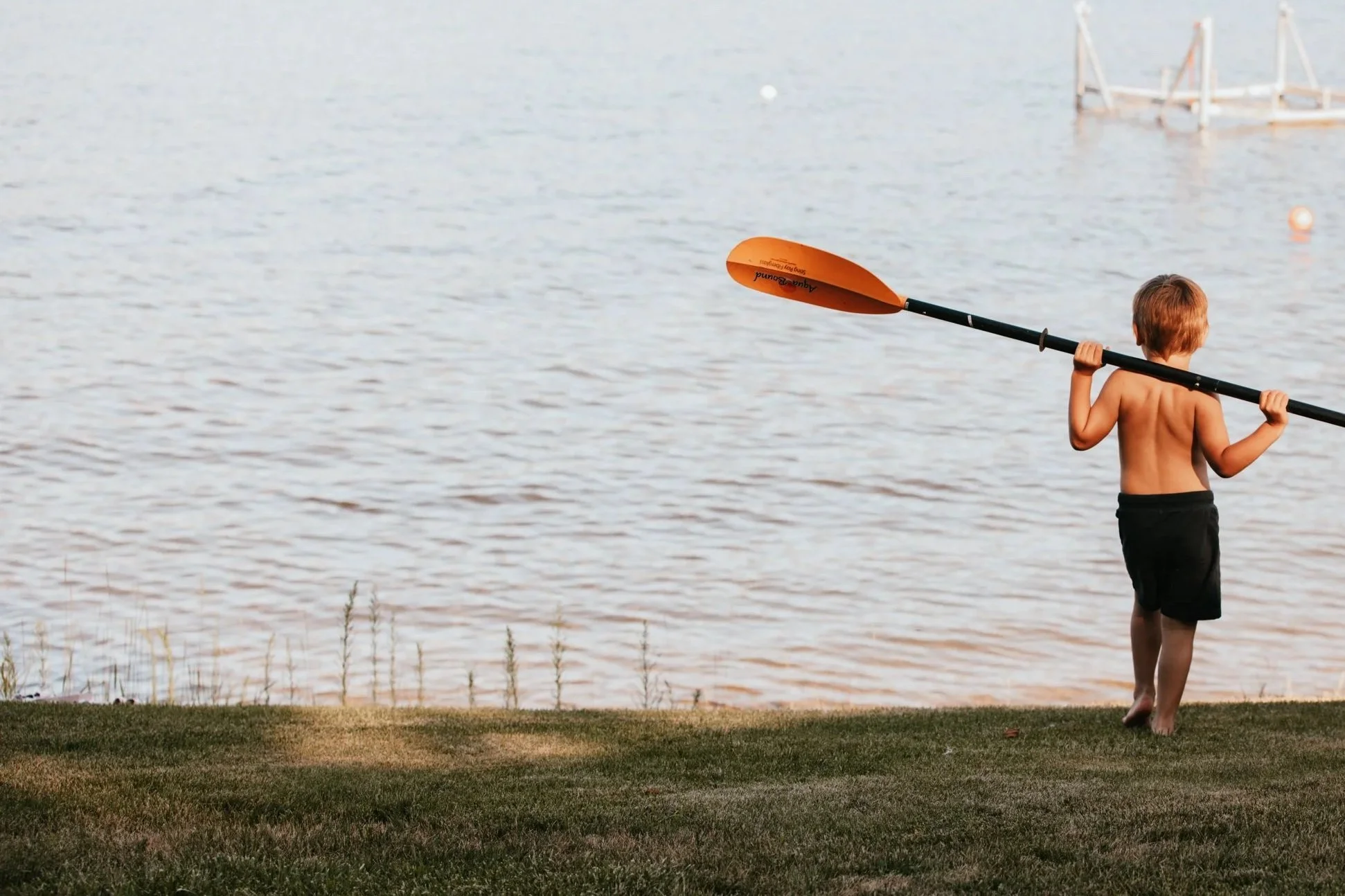 A young boy with no shirt and black shorts holding an orange paddle stands on grass near a lake, facing away, preparing to paddle.