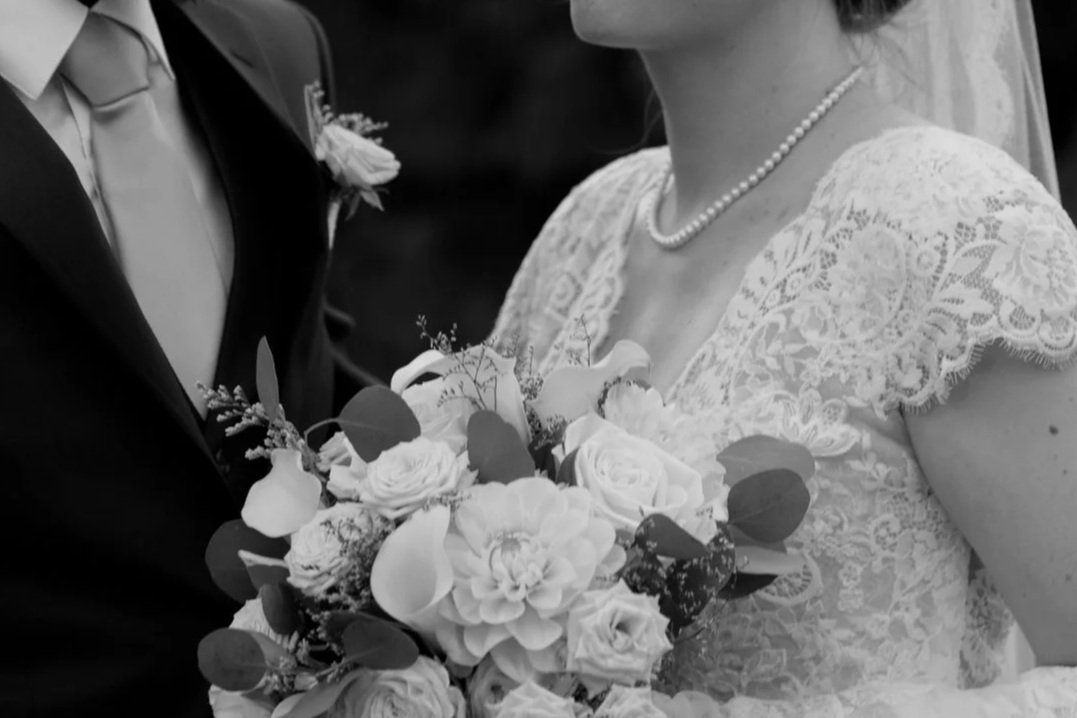 Close-up of a bride holding a bouquet of flowers during a wedding ceremony, with a groom dressed in a suit nearby.