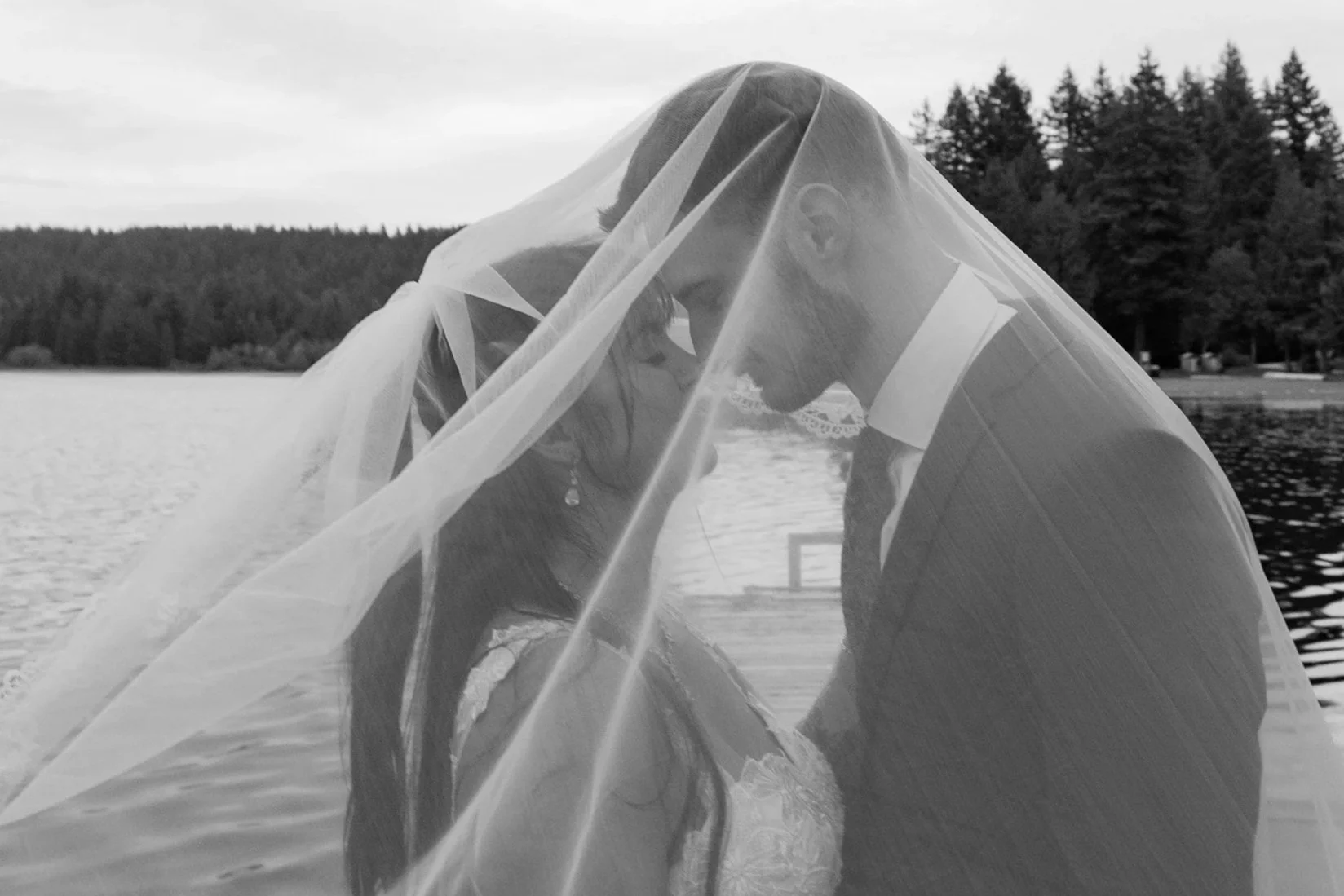 A bride and groom stand close together by a lake, sharing a kiss under the bride's veil, with trees and water in the background.
