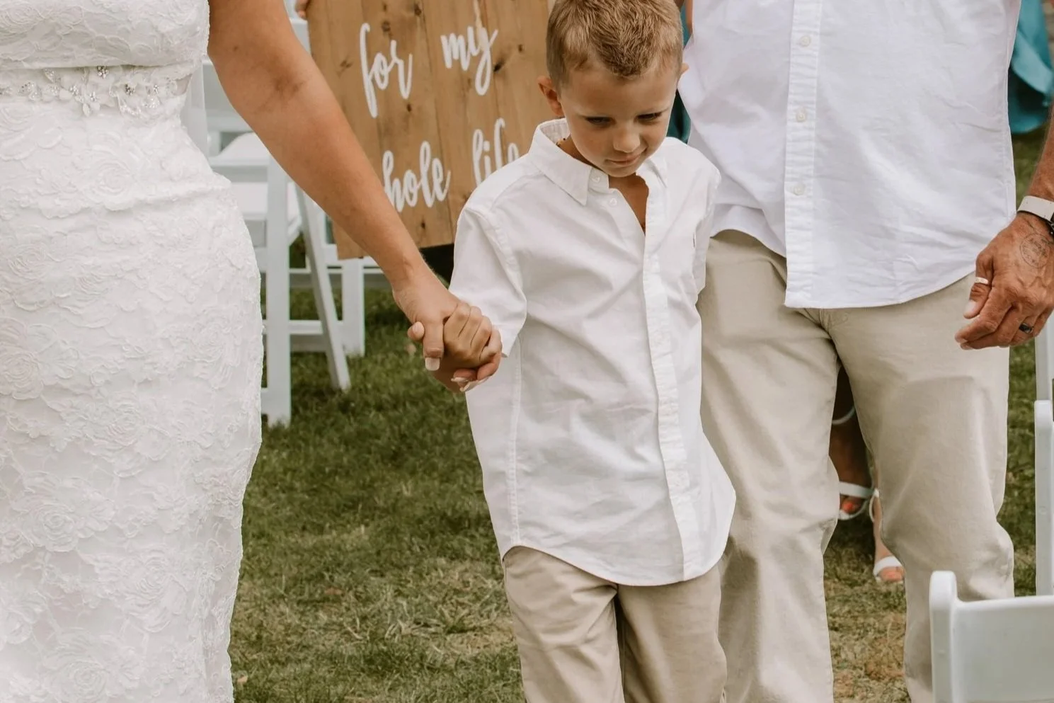 A young boy dressed in a white shirt and tan pants holding hands with an older woman and man, at an outdoor wedding ceremony, with seats and a wooden sign in the background.