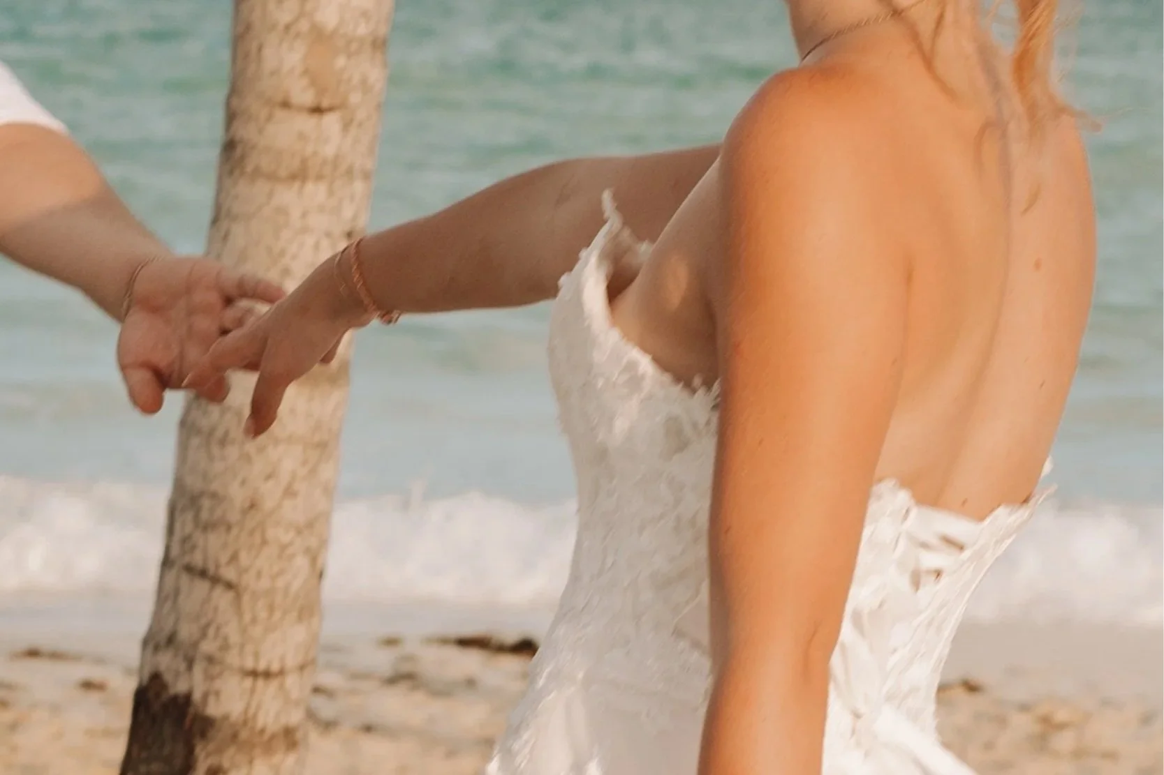 Close-up of a woman in a white dress holding hands with another person at the beach, with the ocean in the background.