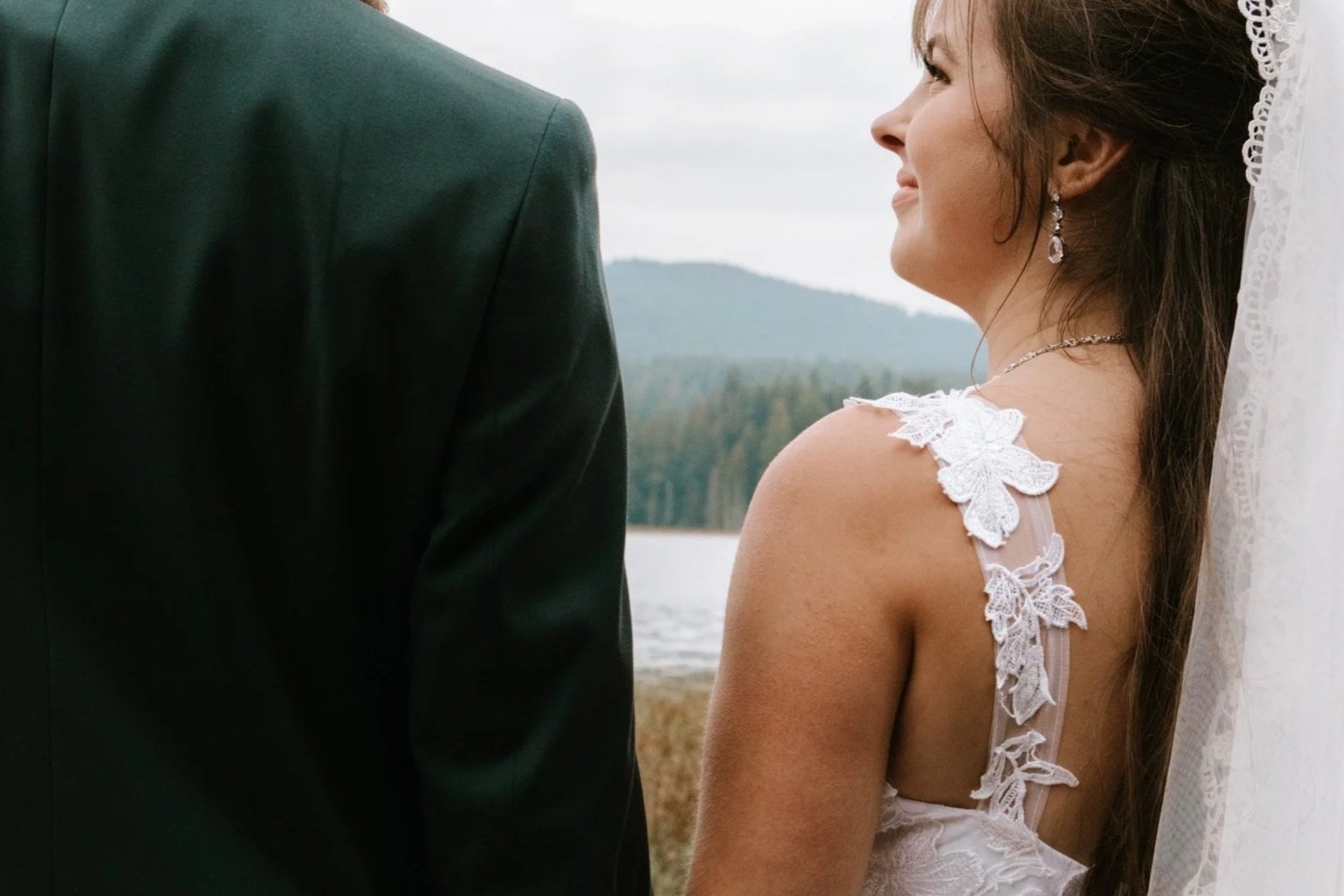 A bride smiling at a groom outdoors near a lake or river with mountains in the background, wearing a wedding dress with lace details and a veil.