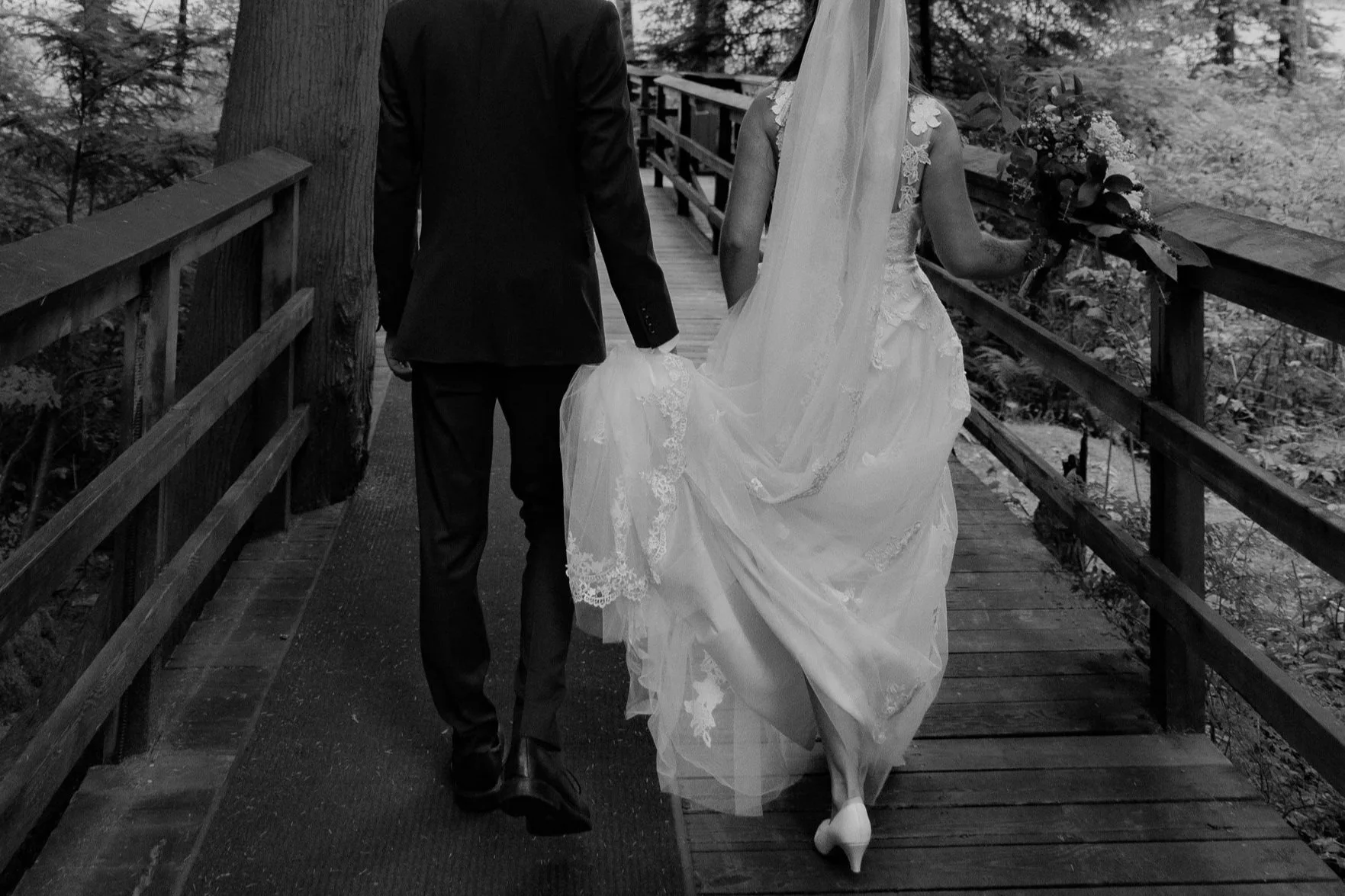 A couple in wedding attire walking hand in hand on a wooden bridge in a wooded area; the bride is holding a bouquet of flowers.