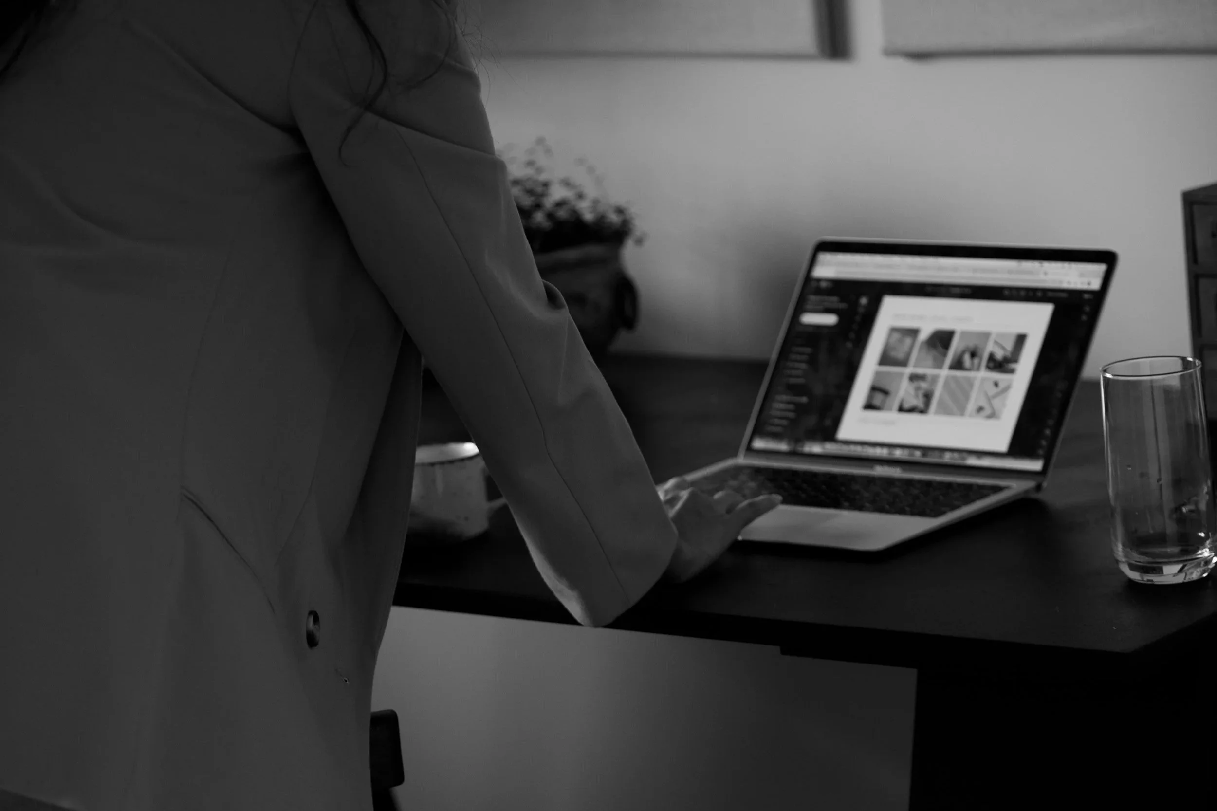A person working on a laptop at a desk, with a glass of water beside it in a monochrome setting.