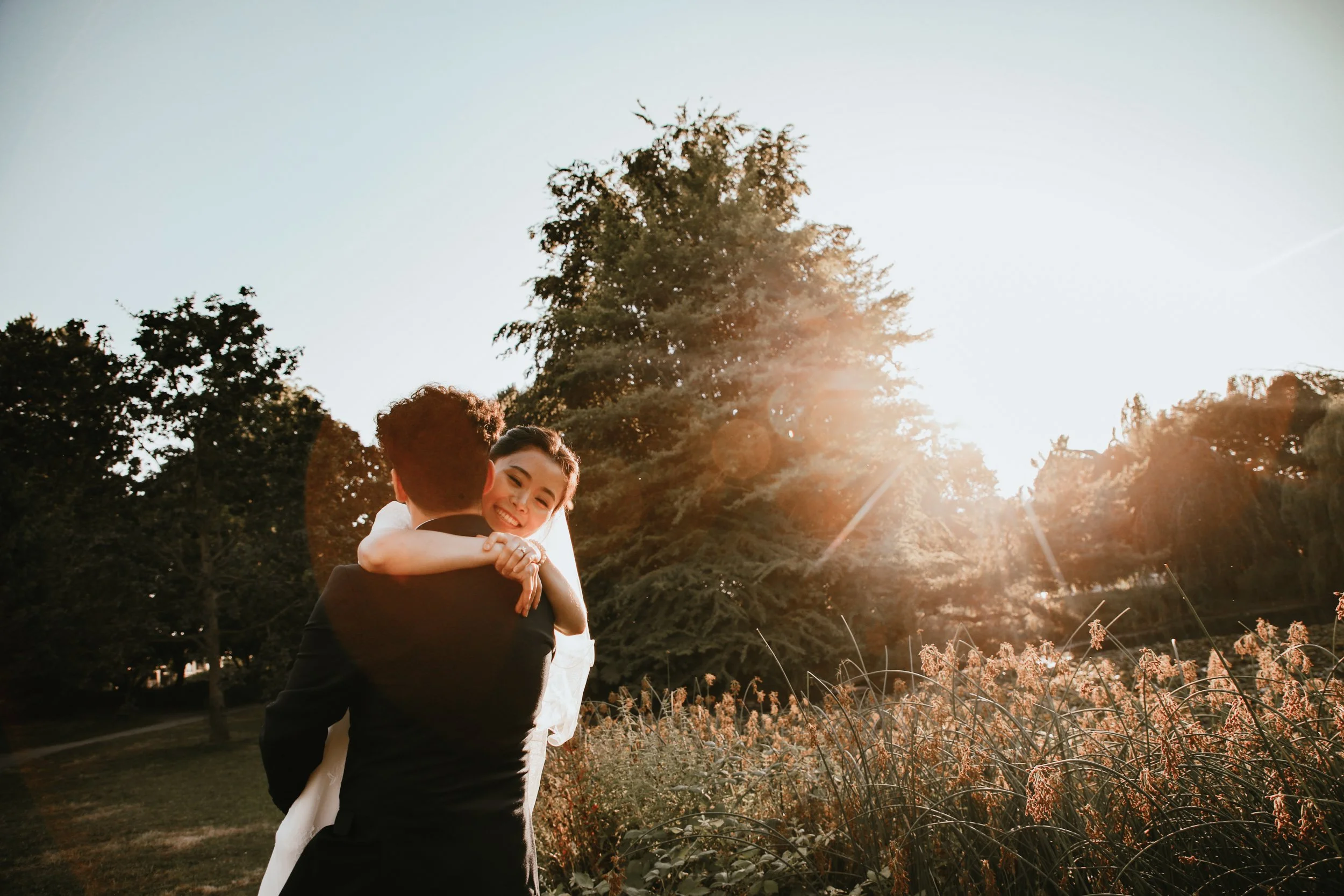 A bride and groom embracing outdoors at sunset, with the bride smiling and the groom facing away, surrounded by trees and tall grass.