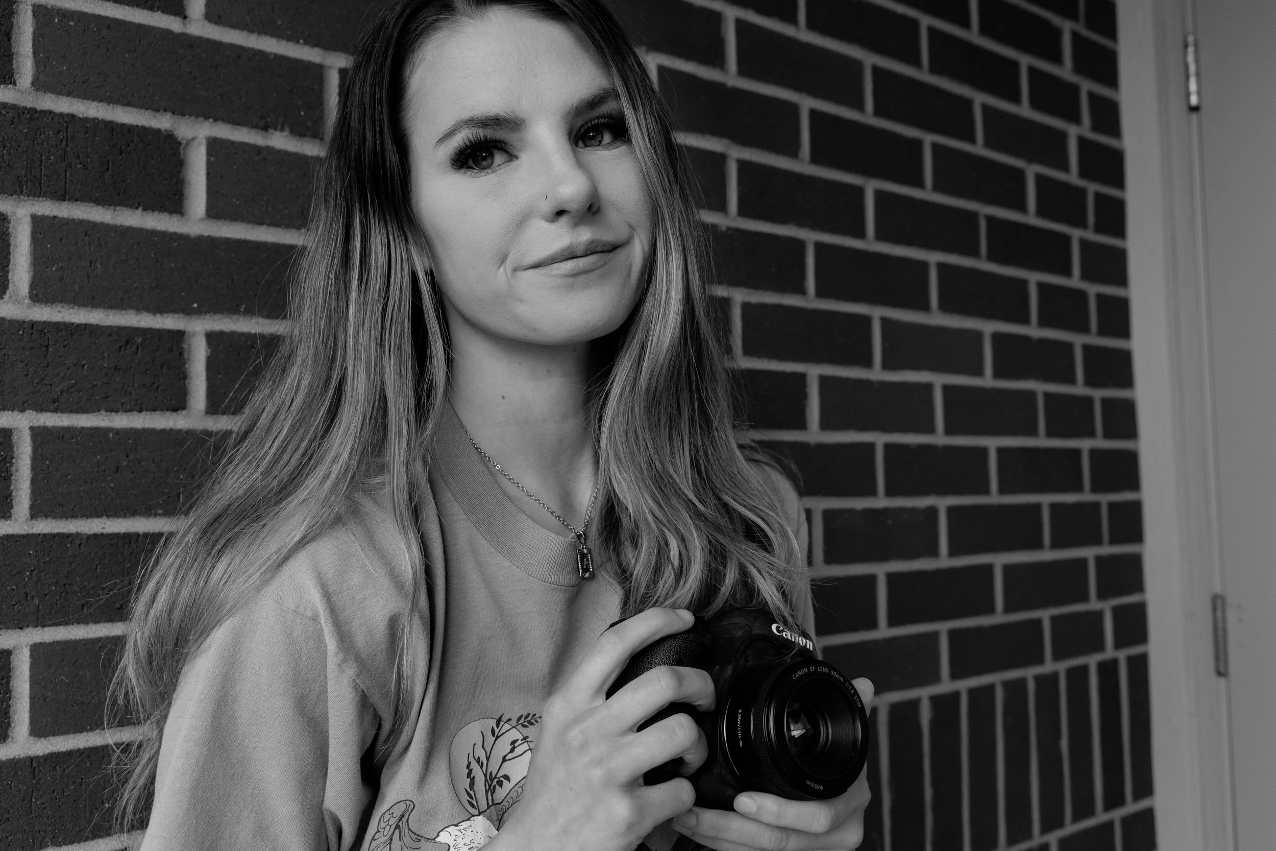 Black and white photo of a young woman with long hair holding a DSLR camera, standing against a brick wall.