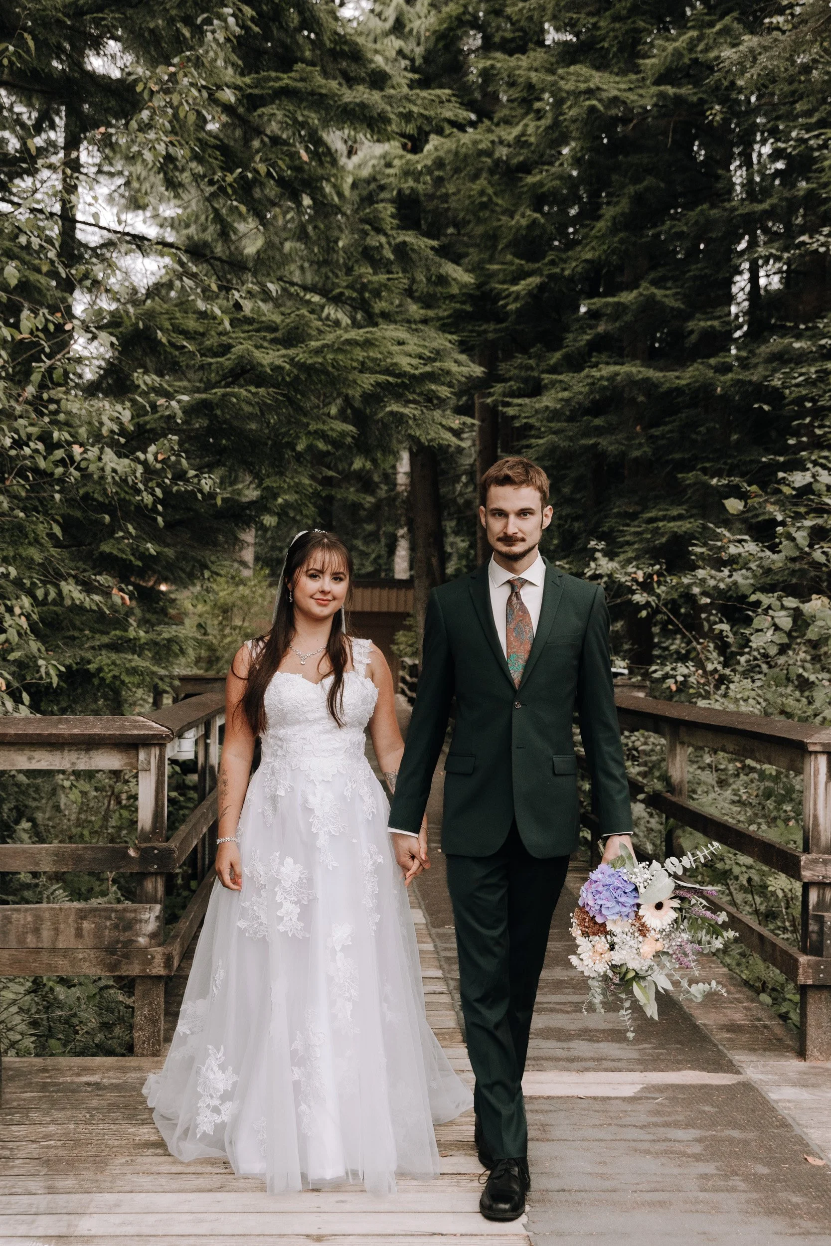 A bride and groom walking hand in hand on a wooden bridge surrounded by tall green trees.