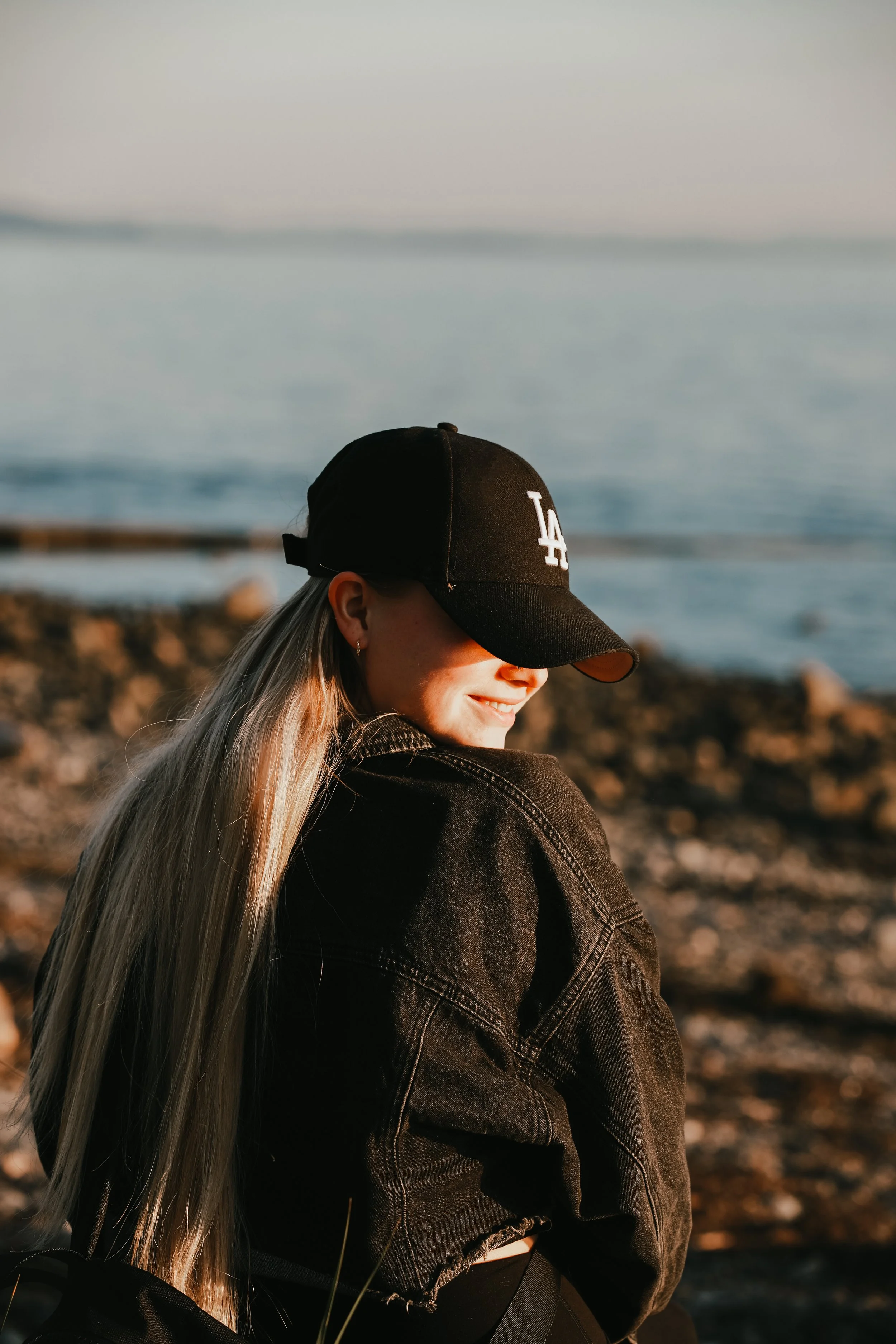 A young woman with blonde hair wearing a black LA baseball cap and a black denim jacket, sitting on the rocky shore near the ocean, smiling and looking down.
