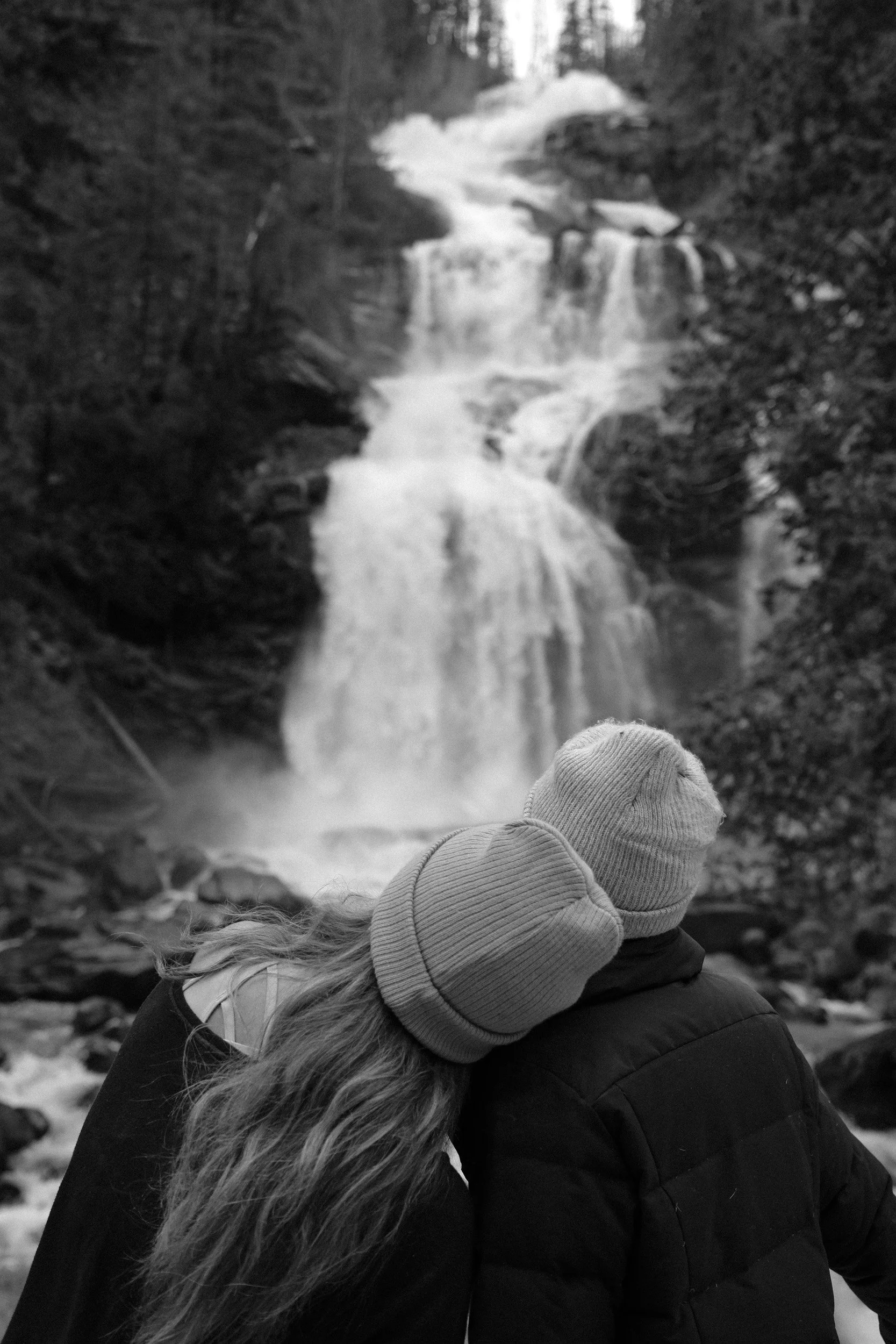 A black-and-white photo of a couple wearing knit caps, sitting close together and looking at a waterfall in a forested area.