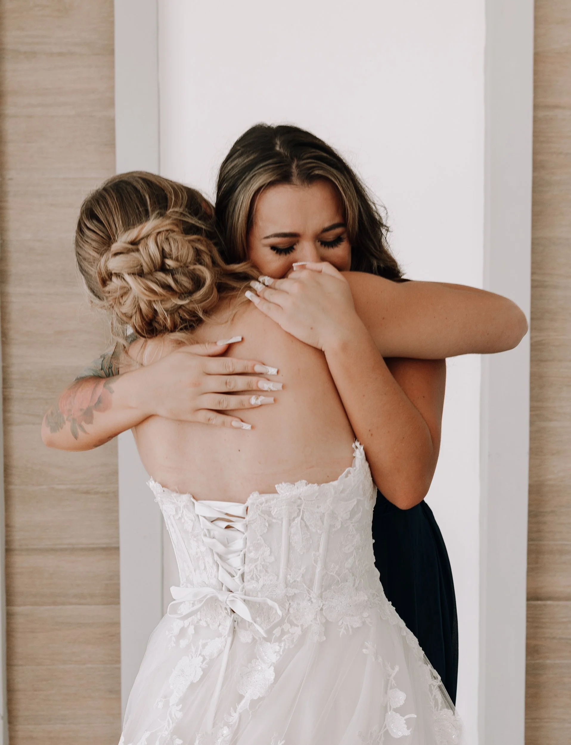 Two women hugging, one in a wedding dress, the other in a dark dress, emotionally embracing in a heartfelt moment.