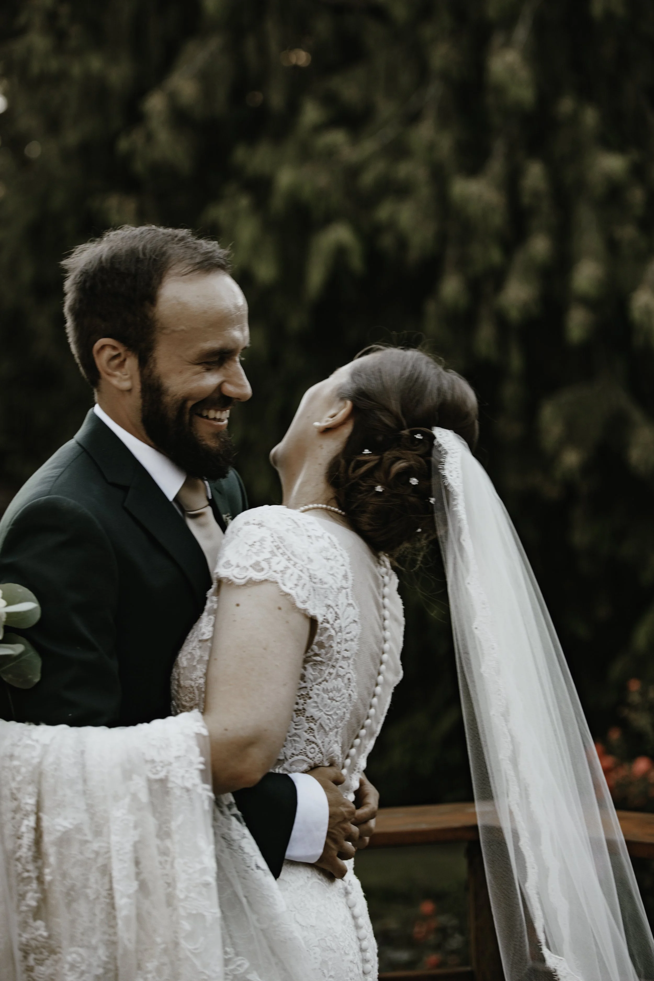 Bride and groom share a joyful moment outdoors during their wedding, with trees and blurred greenery in the background.