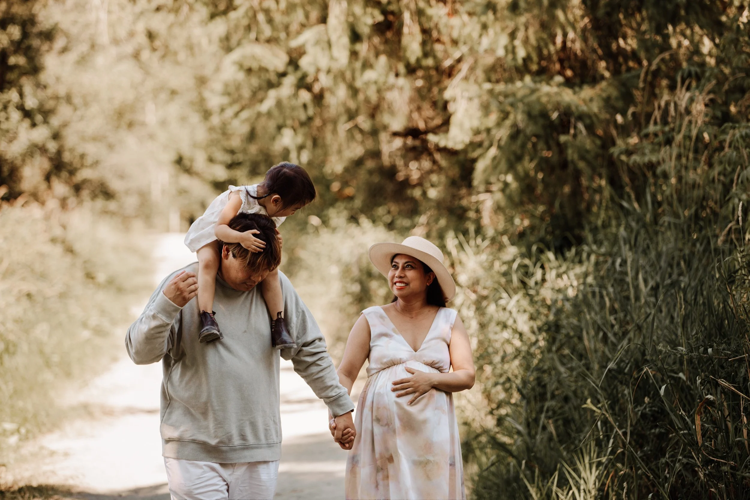 A family of three walking outdoors on a sunny day in a lush green forest. The father is carrying a young girl on his shoulders, holding her hand, while the mother, dressed in a flowing dress and wide-brimmed hat, walks beside them looking at the girl