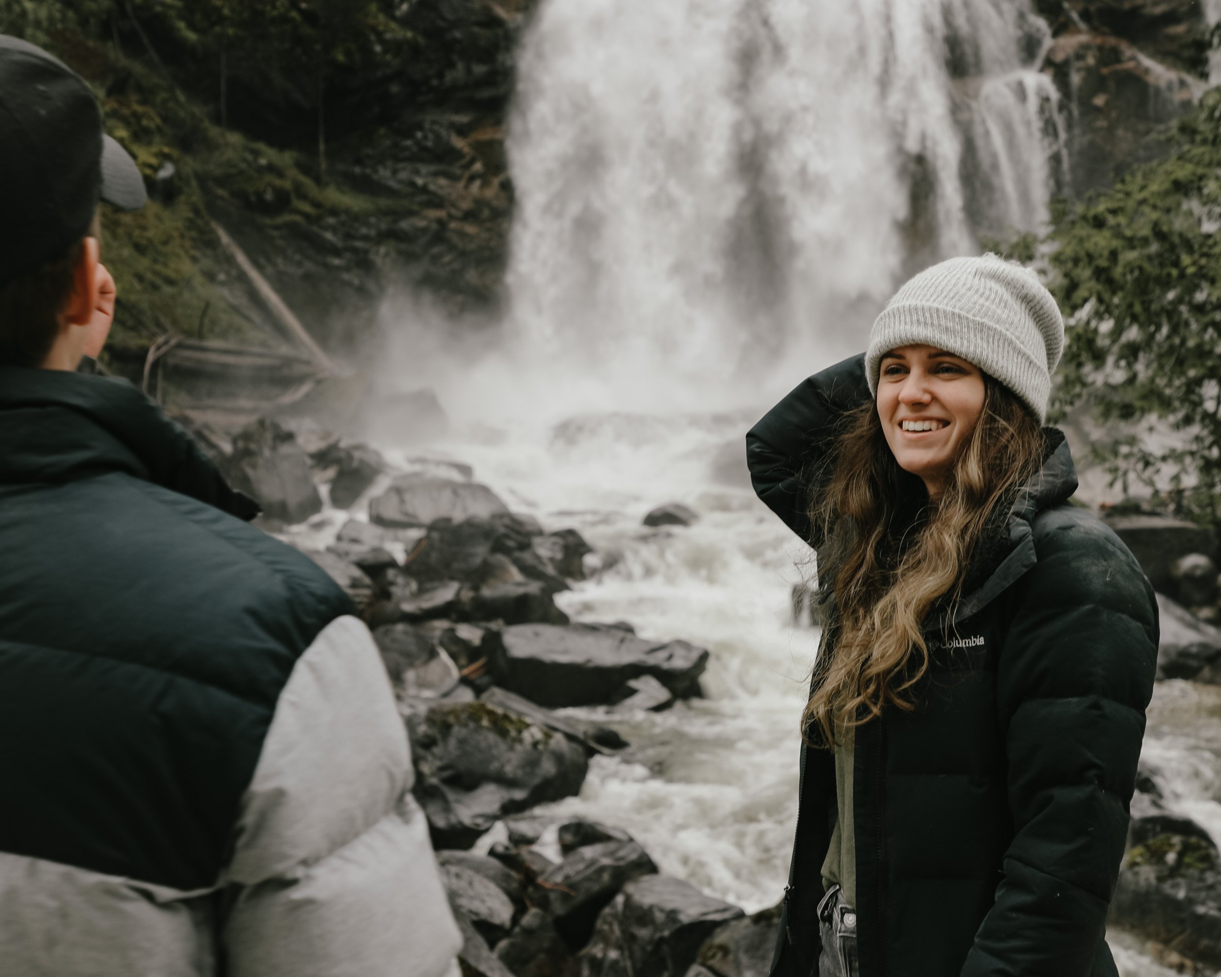 A young woman wearing a grey beanie and black Columbia jacket smiling and adjusting her beanie near a waterfall with rocks and rushing water in the background.