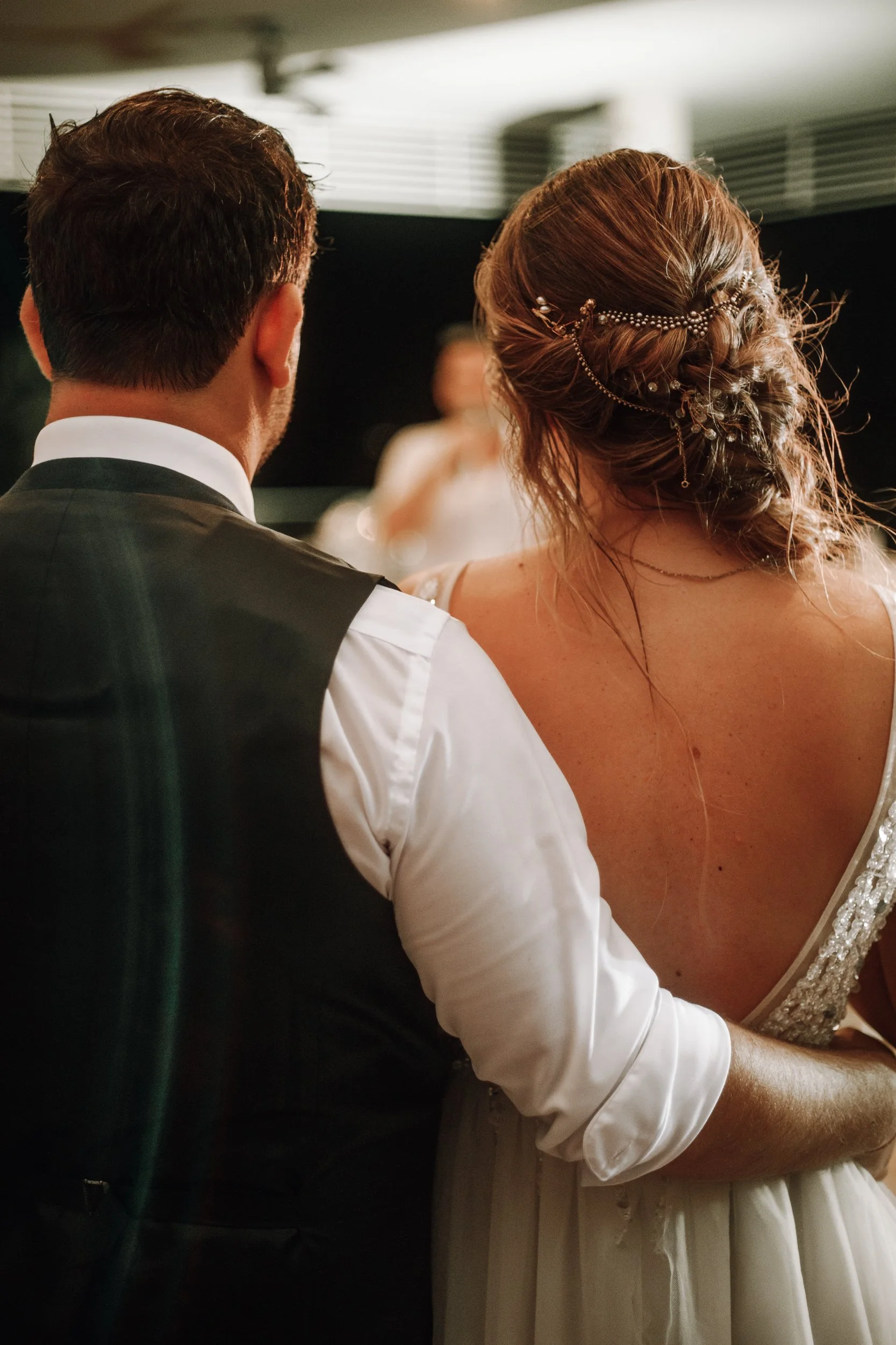 Back view of a man and a woman at a wedding ceremony, with the man wearing a black vest and white shirt, and the woman in a white dress with intricate braid hairstyle and decorative hair accessories.
