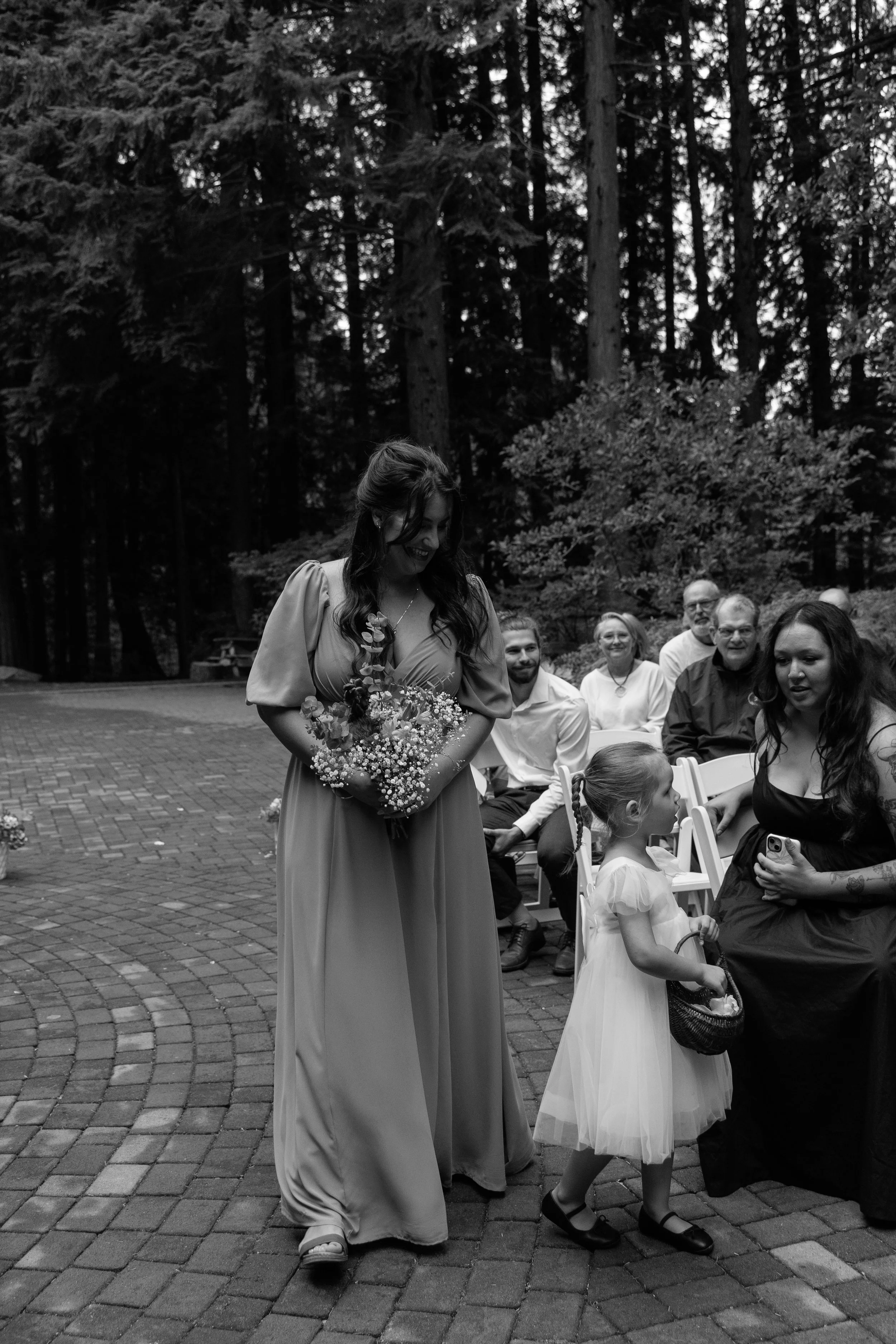 A woman in a long dress holding a bouquet of flowers, walking past a young girl dressed in a fancy dress holding a basket, as seated guests watch during an outdoor wedding ceremony in a wooded area.