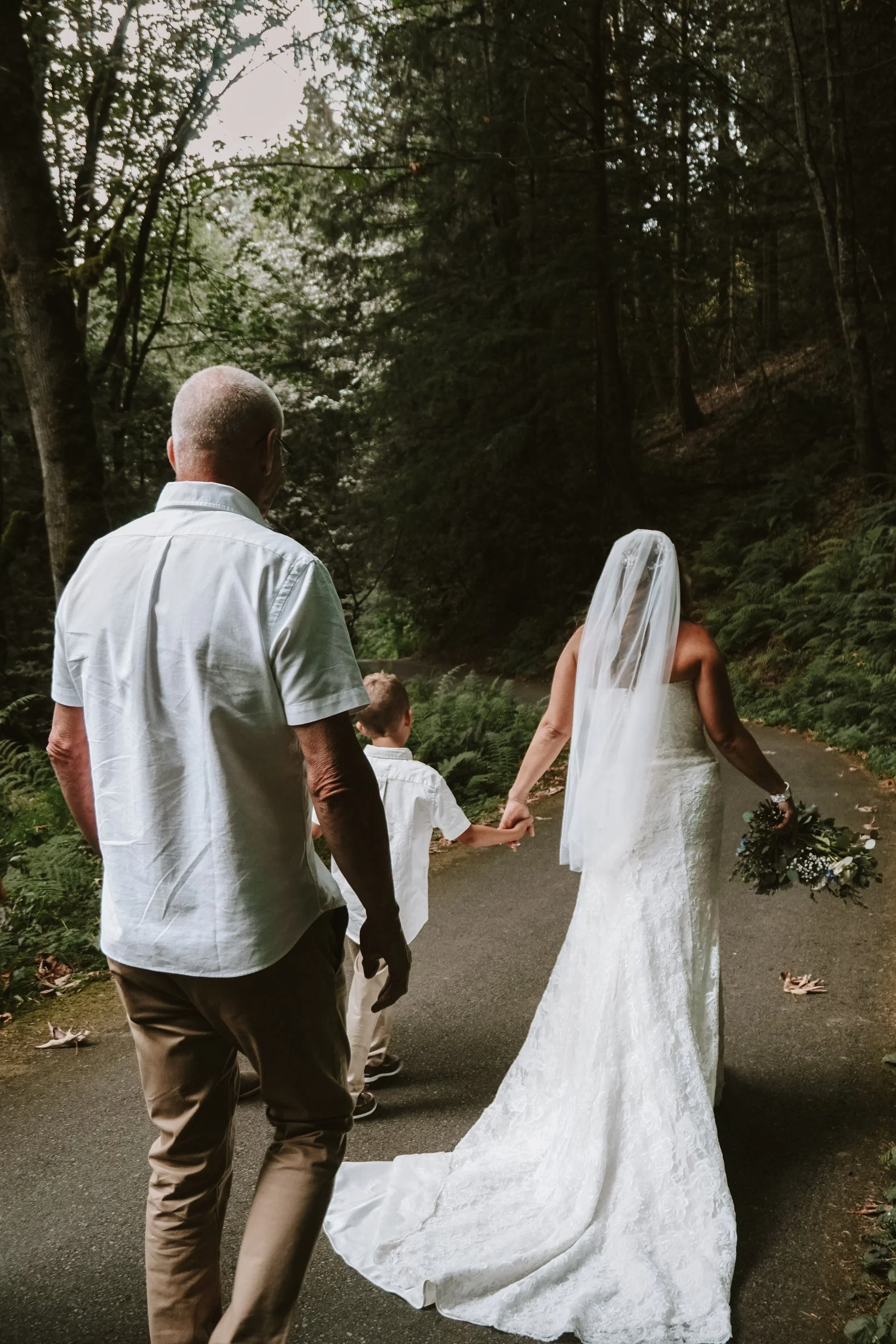 A bride in a white lace wedding dress with a veil holding a bouquet, walking hand-in-hand with a woman, possibly her mother, on a forest path, accompanied by a man and a young boy.