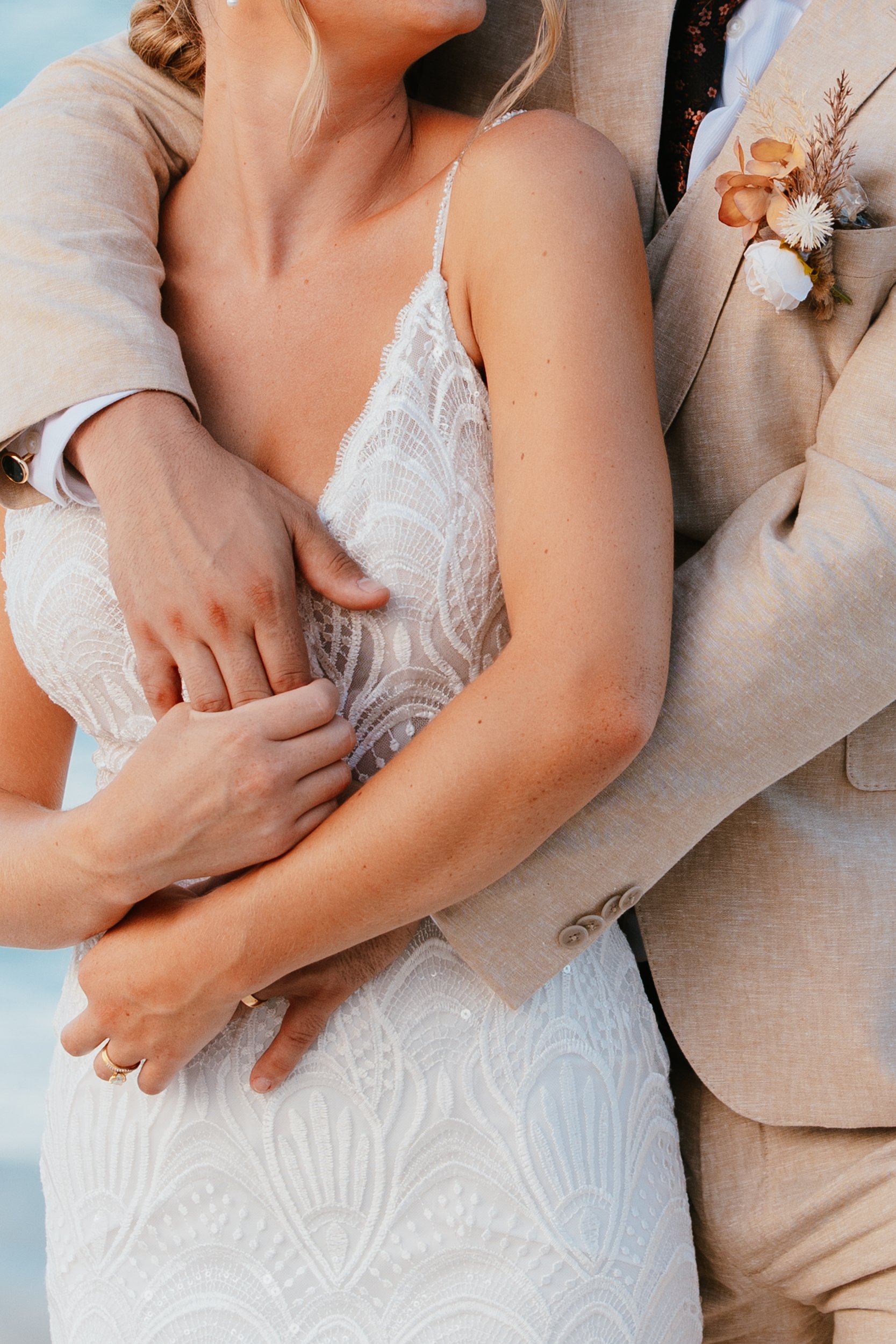 Close-up of a newlywed couple embracing, showing the bride's lace wedding dress and the groom in a beige suit with a boutonniere, with their hands intertwined.