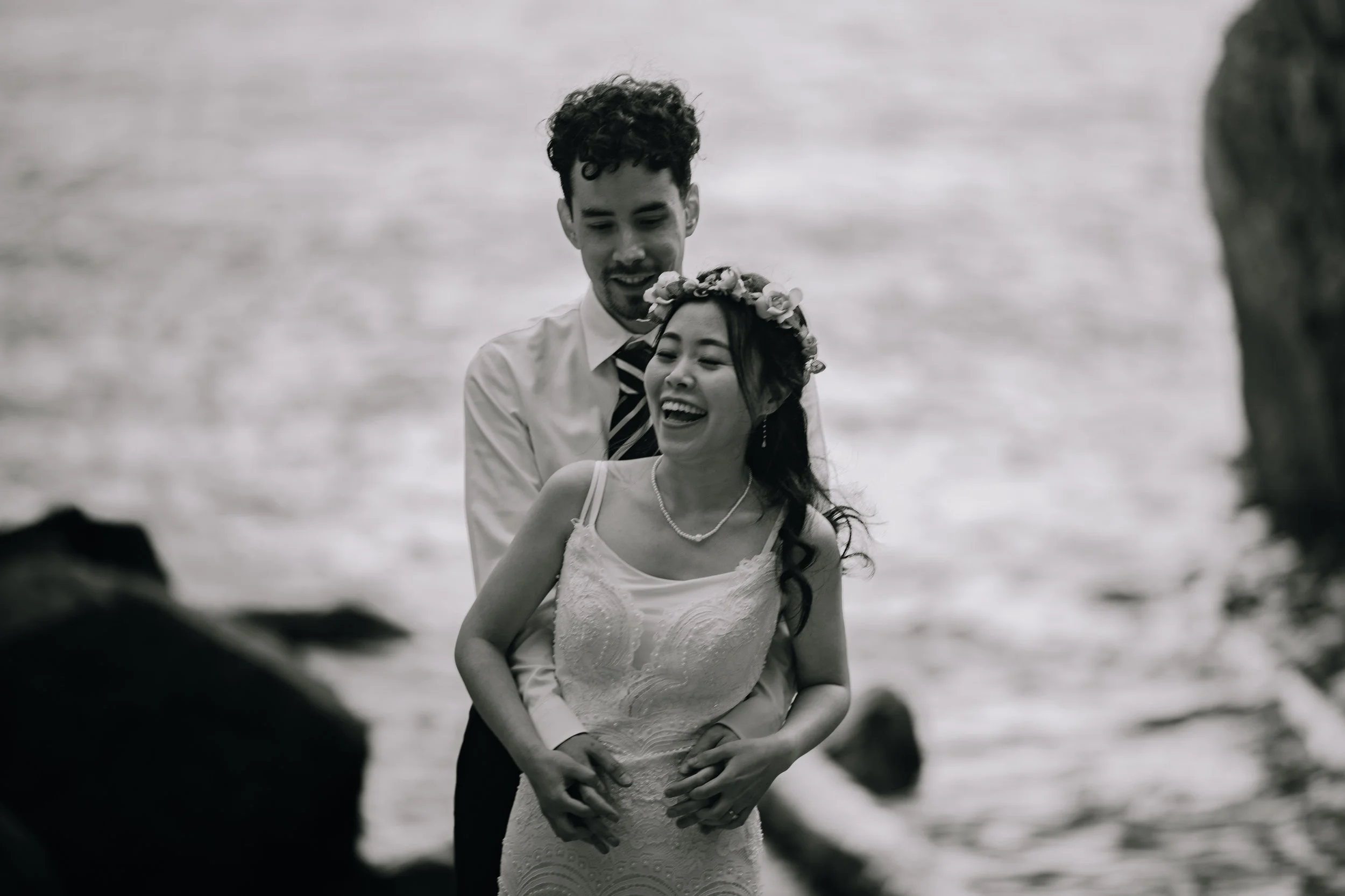 A joyful couple in wedding attire, standing on a rocky shoreline with the ocean in the background, sharing a happy moment.
