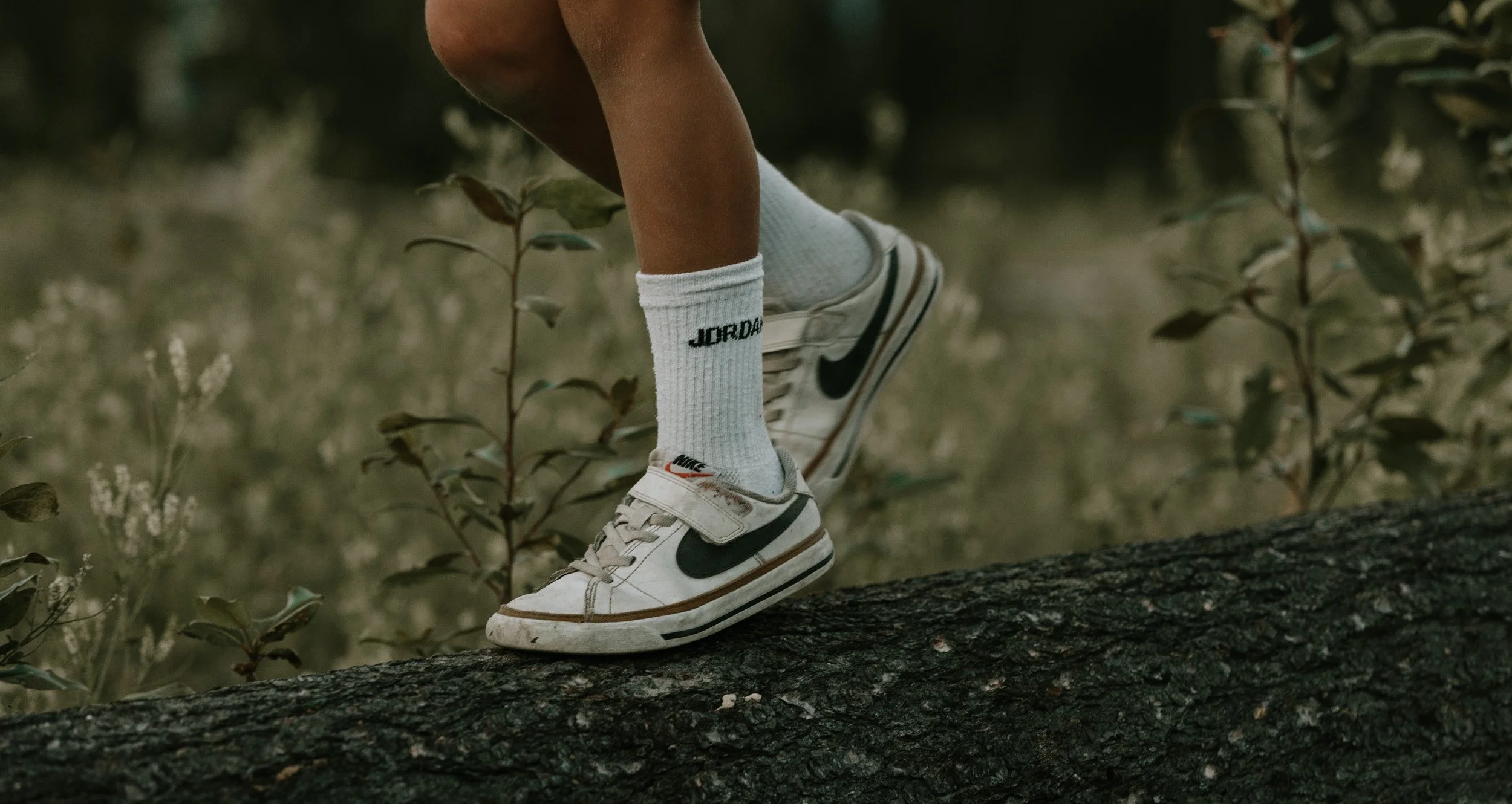 A close-up of a person's feet and legs balancing on a fallen tree log in a natural setting, wearing white Nike sneakers and white socks with black text.