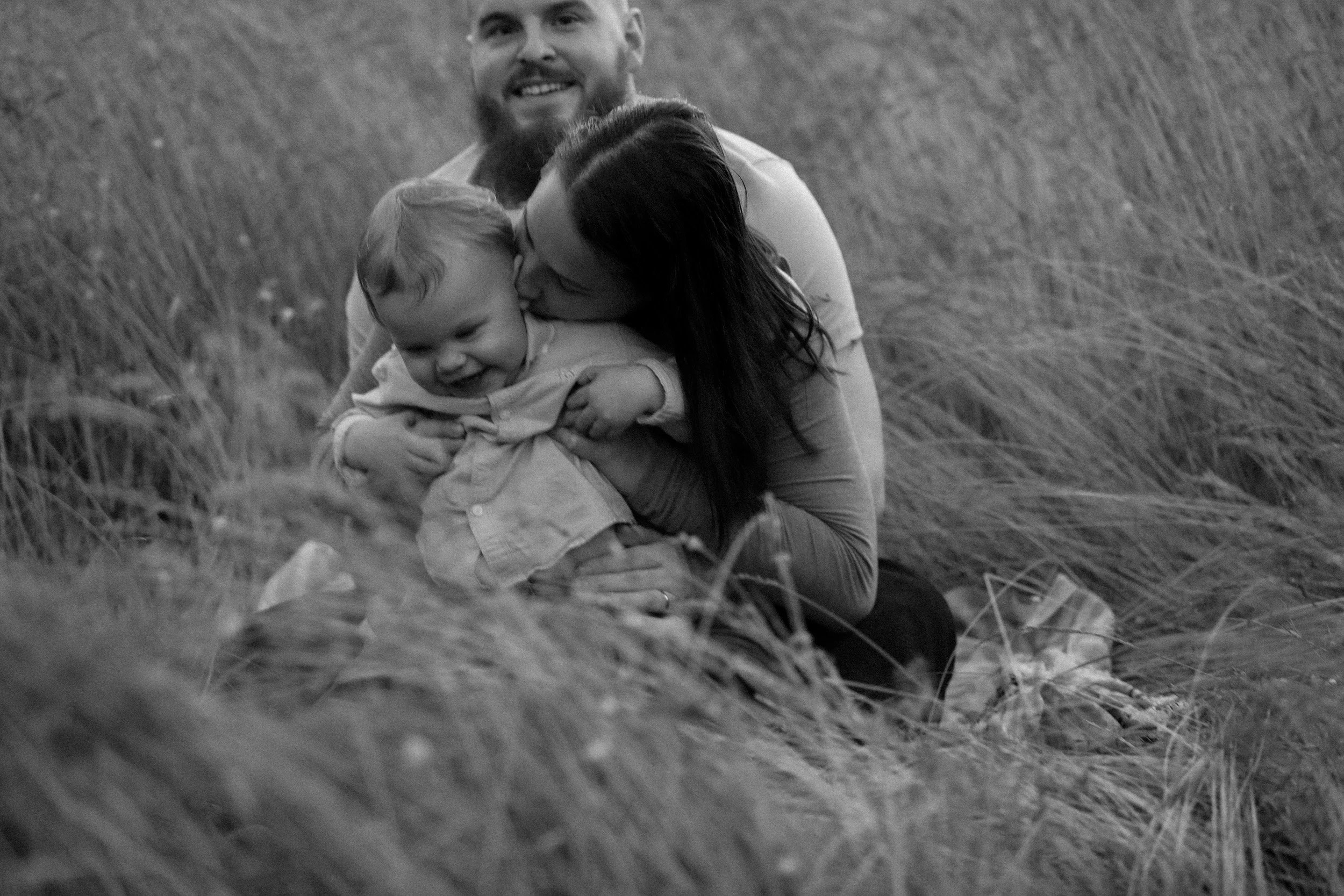 A family of three, a man, woman, and young child, sitting on the ground in a grassy outdoor area, enjoying a moment of affection and closeness.