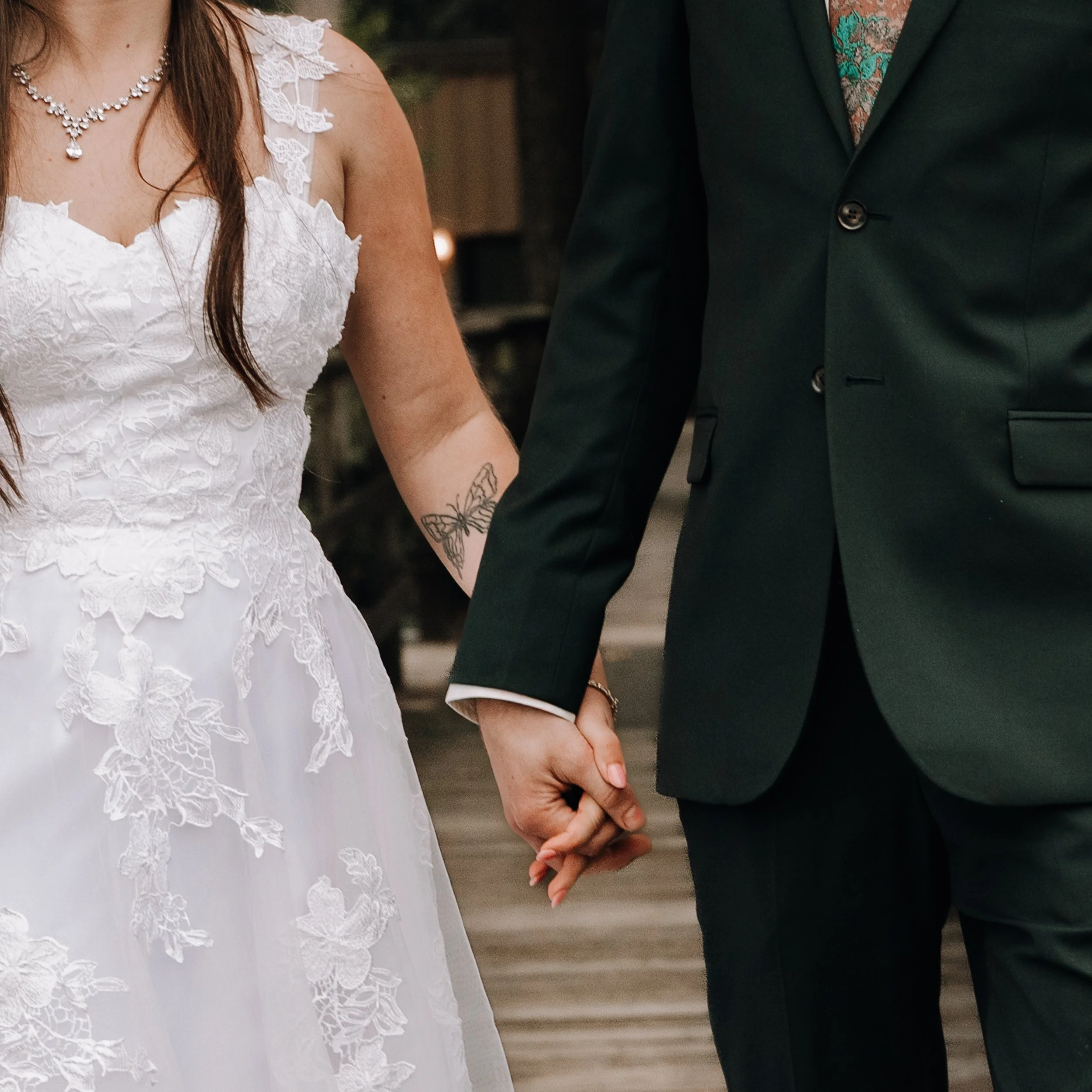 A bride and groom holding hands at their wedding, with the bride wearing a white lace dress and the groom wearing a dark suit.