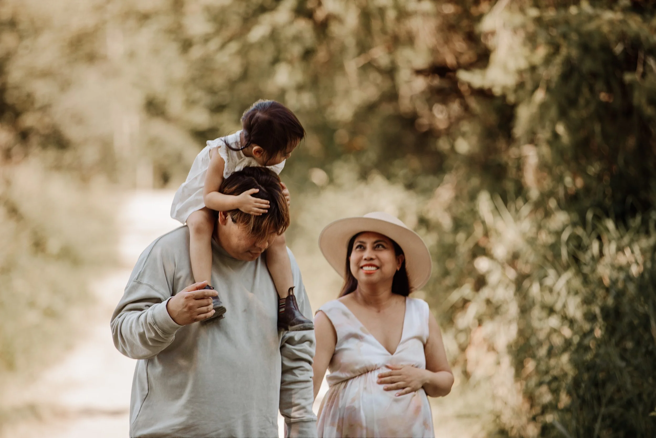 A family of three walking outdoors in a forested area with a dirt path. The father carries a young girl on his shoulders, who is hugging his head. The mother walks beside them, smiling and holding her belly, wearing a large sun hat.
