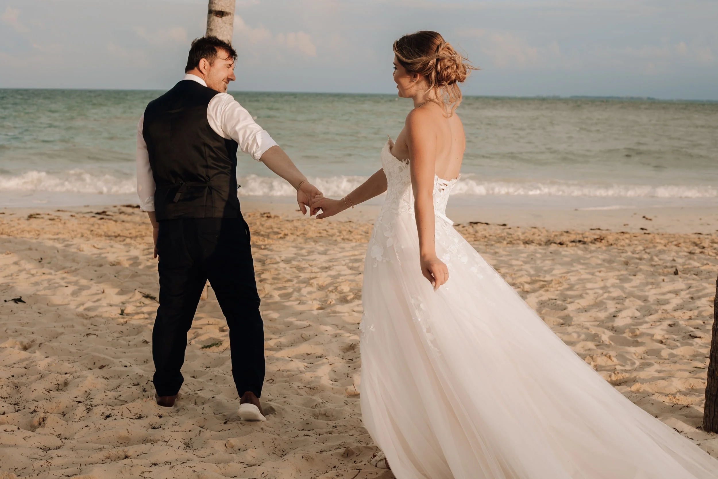 A newlywed couple holding hands on a beach, with the ocean in the background, the woman in a white wedding dress and the man in a vest and dress pants.