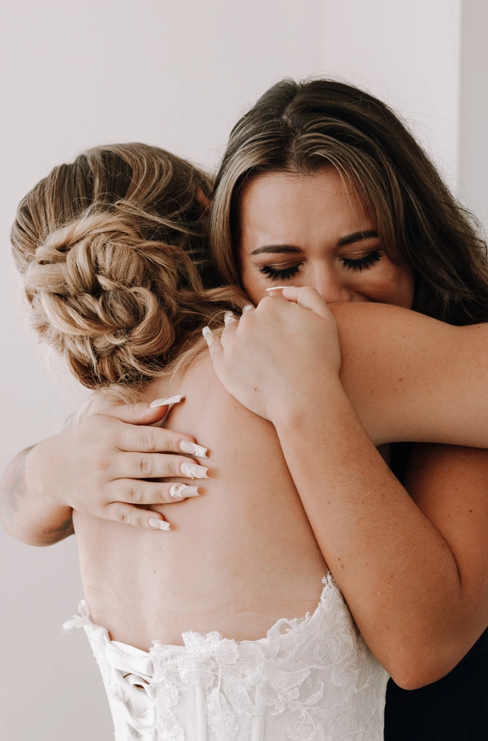 Two women sharing an emotional hug, one in a white lace wedding dress, the other wearing a black top, in an intimate moment.