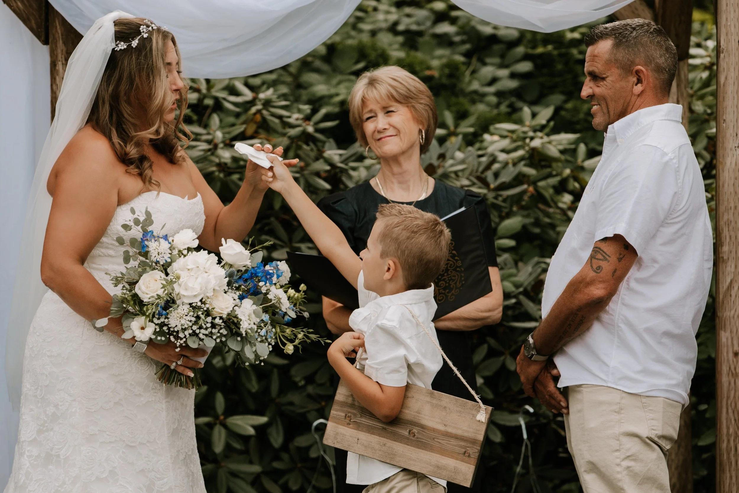 A bride in a white wedding dress holding a bouquet, exchanging rings with a groom in a white shirt, during a wedding ceremony outdoors, with a young boy dressed in white and a black woman officiating in the background.