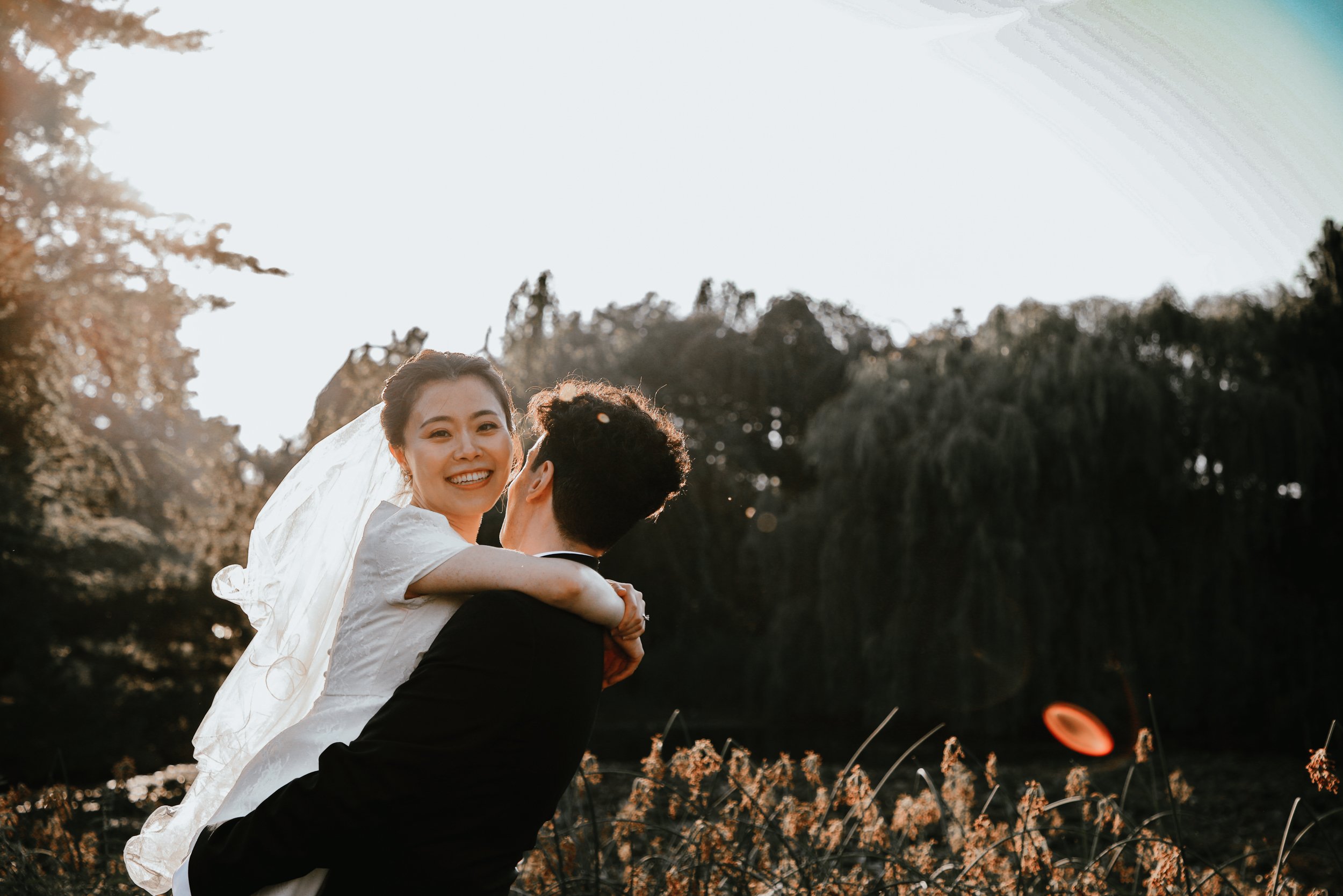 A joyful bride in a white wedding dress and veil being lifted by a groom in a black suit outdoors during sunset, with trees and greenery in the background.