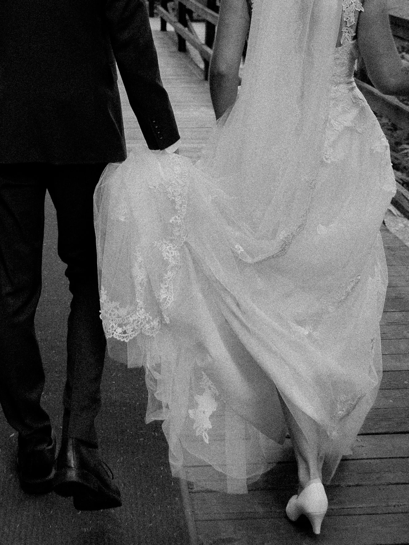 A bride in a wedding dress and a groom in a suit holding hands as they walk on a wooden bridge.