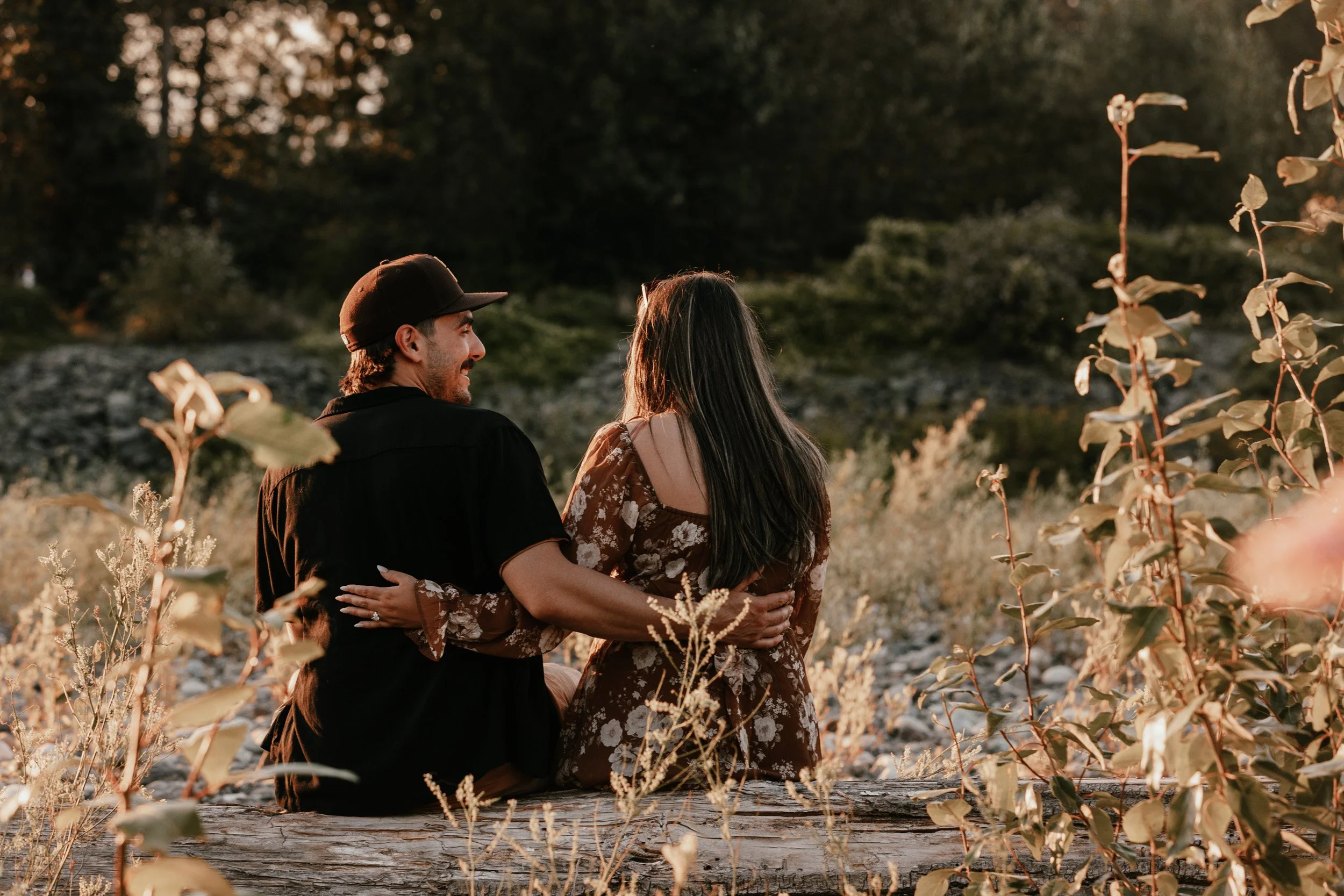 A couple sitting on a log in a natural outdoor setting, embracing and looking at each other during sunset, with trees and bushes in the background.