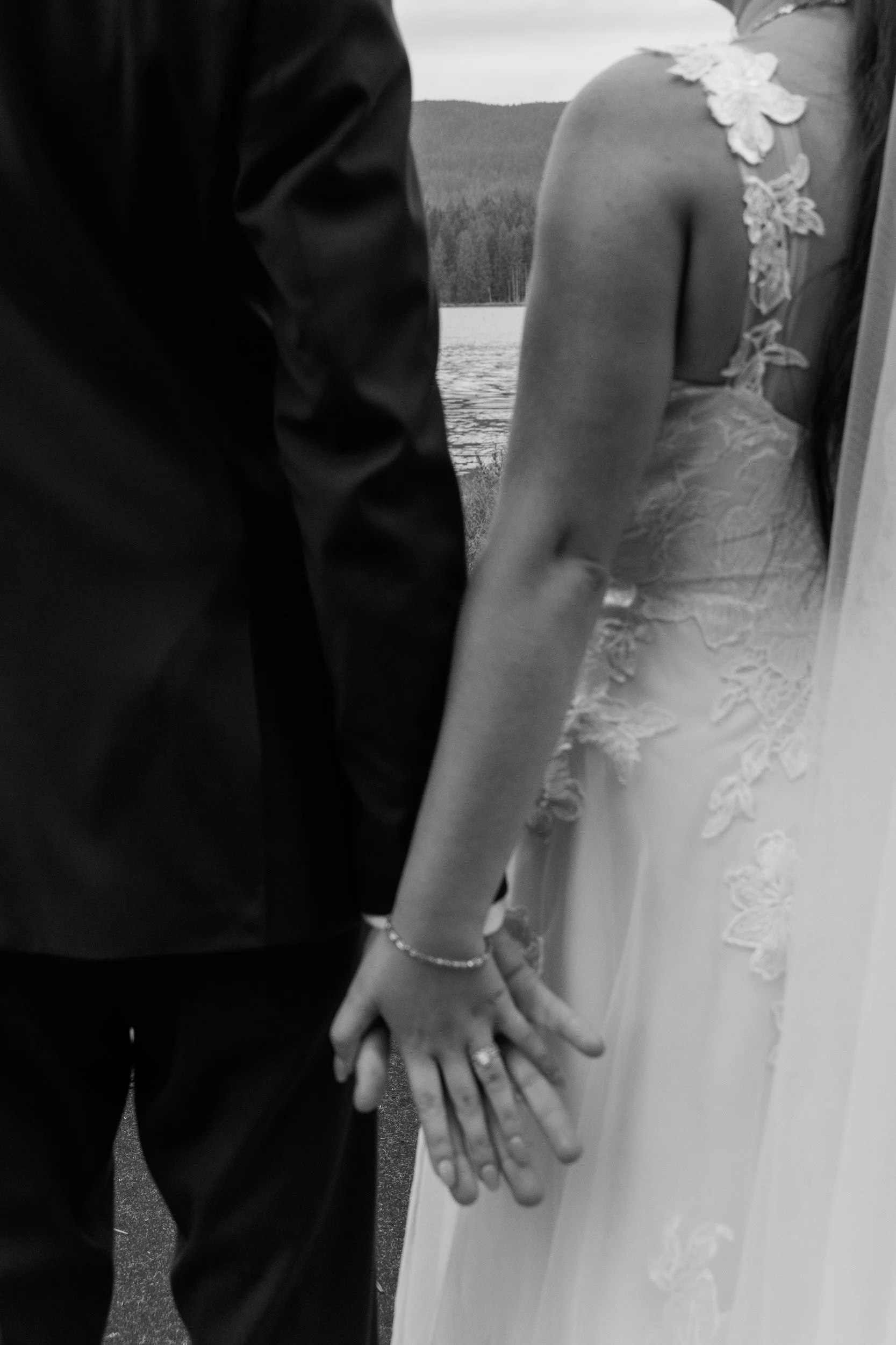 A bride and groom holding hands, with the bride wearing a lace wedding dress and the groom in a dark suit, standing outdoors near a lake and forested area.