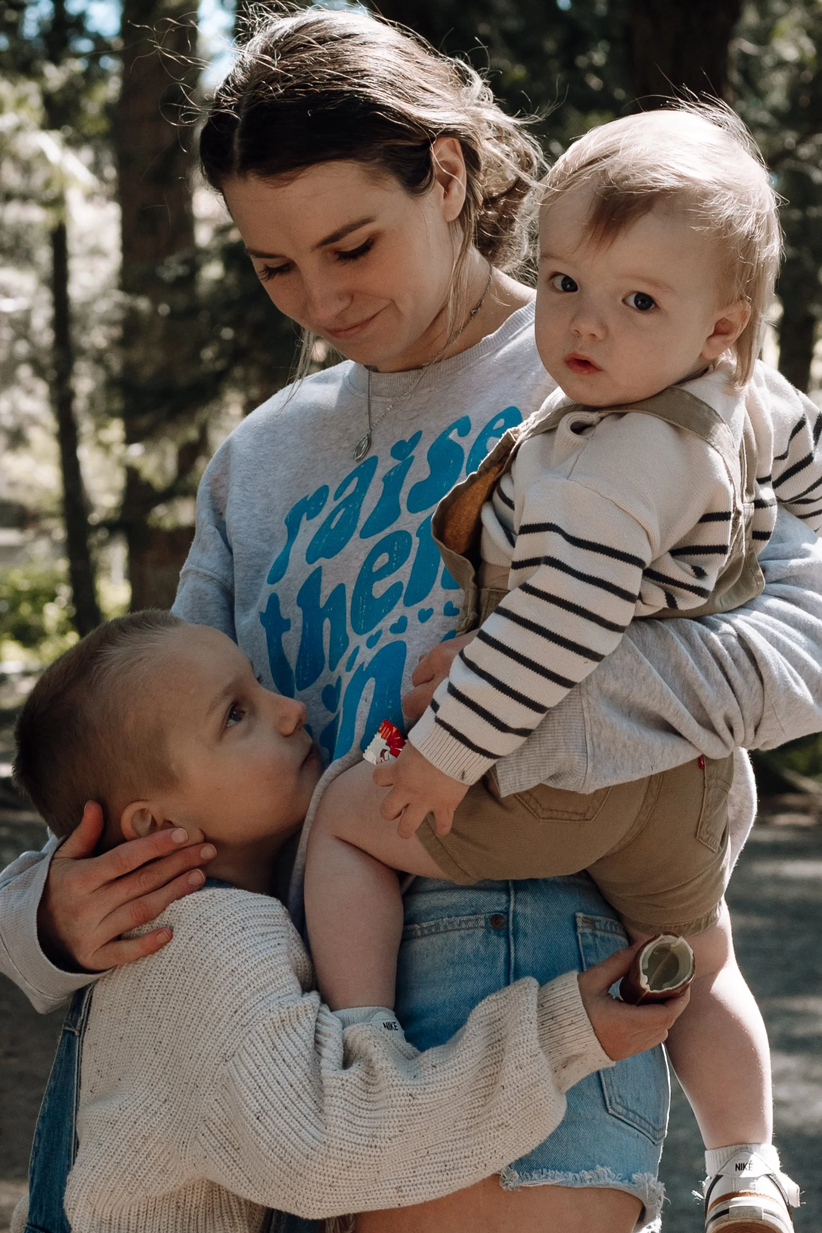 A young woman holding two children outdoors in a wooded area, one child resting on her shoulder and the other looking at her while she embraces him; the scene depicts a tender moment.