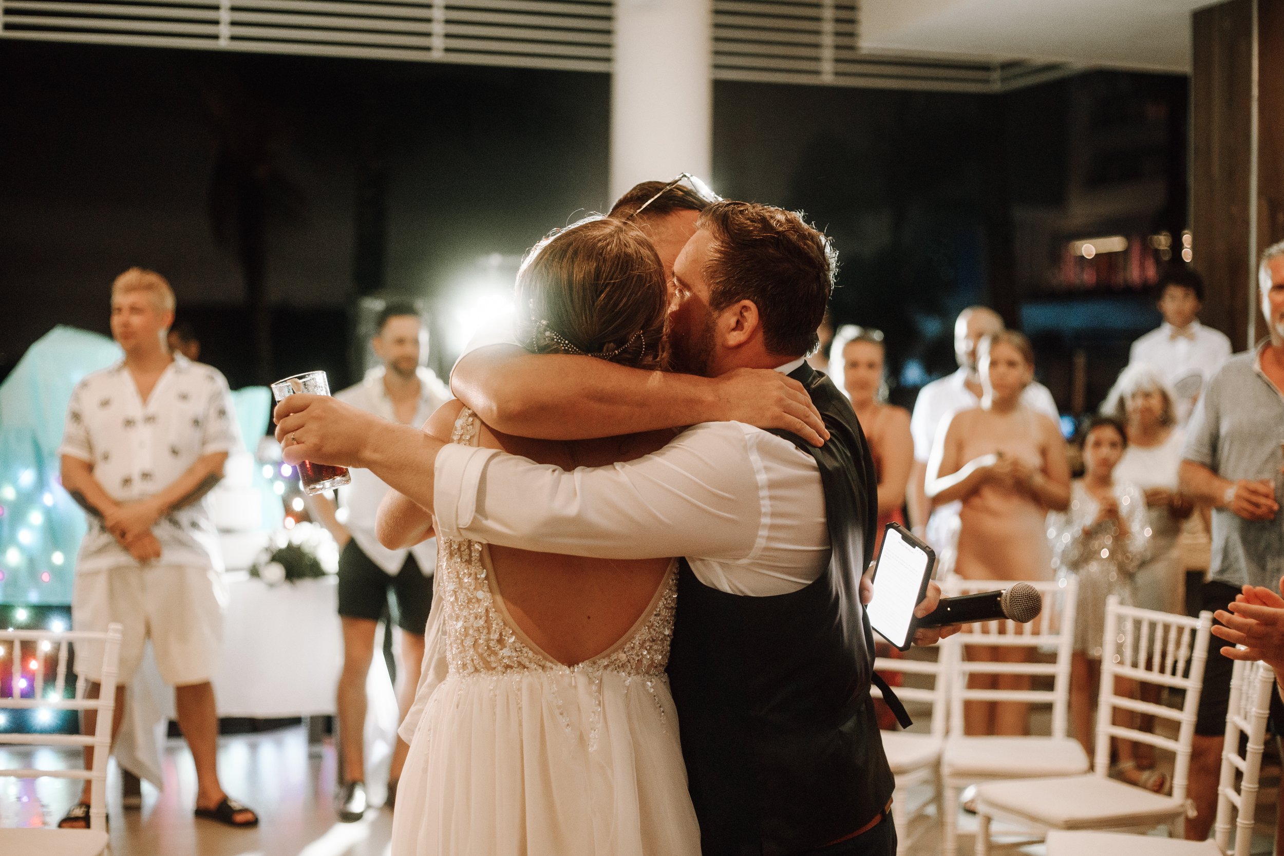 A bride and groom embrace and kiss during their wedding reception, surrounded by guests in an indoor venue with chairs and festive decorations.