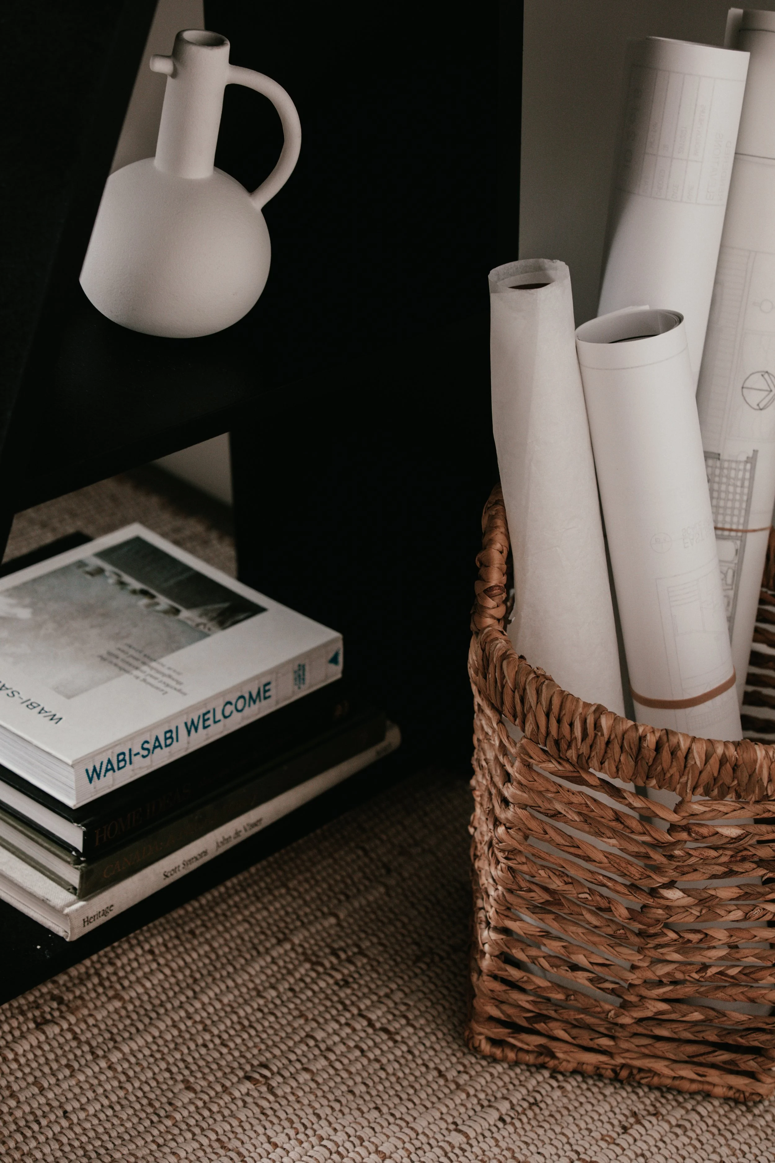 A black shelf with a white textured ceramic pitcher on top. Below, there's a stack of magazines or books, including one titled 'WABI-SABI WELCOME.' To the right, a woven basket contains rolled-up papers and blueprints. The surface has a woven, natura