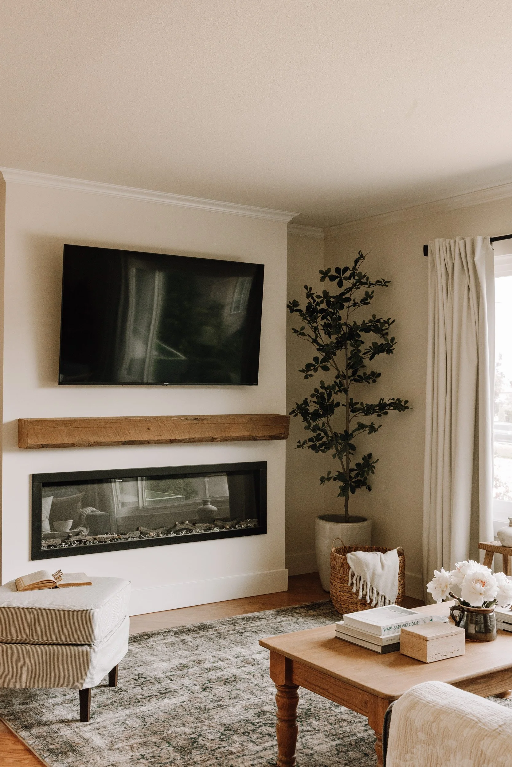 Living room with a wall-mounted flat-screen TV above a wooden mantel, a modern fireplace below the TV, a large potted plant to the right, a cream-colored ottoman, a wooden coffee table with books and flowers, and cream curtains covering a window.