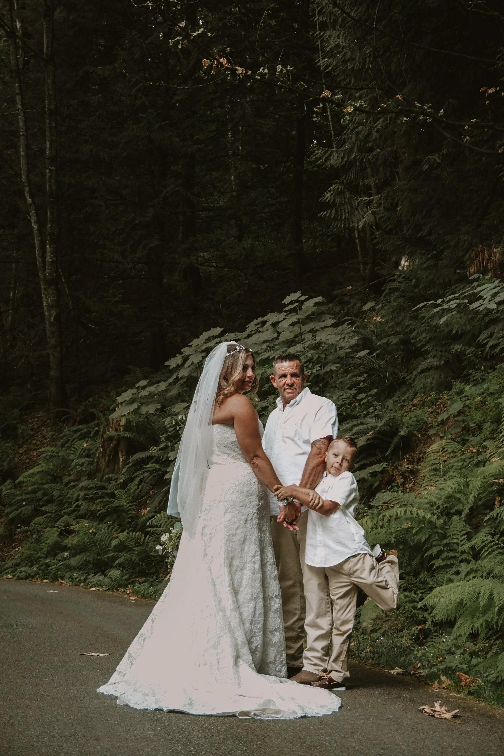 A bride in a white lace wedding dress and veil holding hands with a man and a boy, standing on a wooded road in a forest.