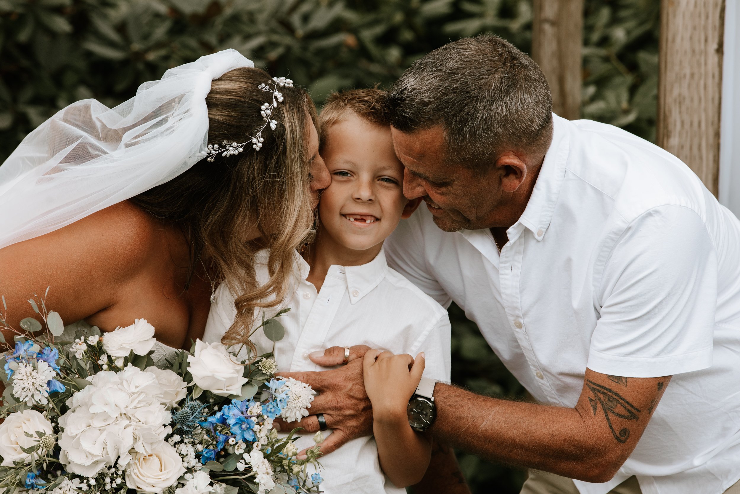 Family celebrating a wedding, with a bride, groom, and a young boy in white shirts, smiling and sharing a moment outdoors.