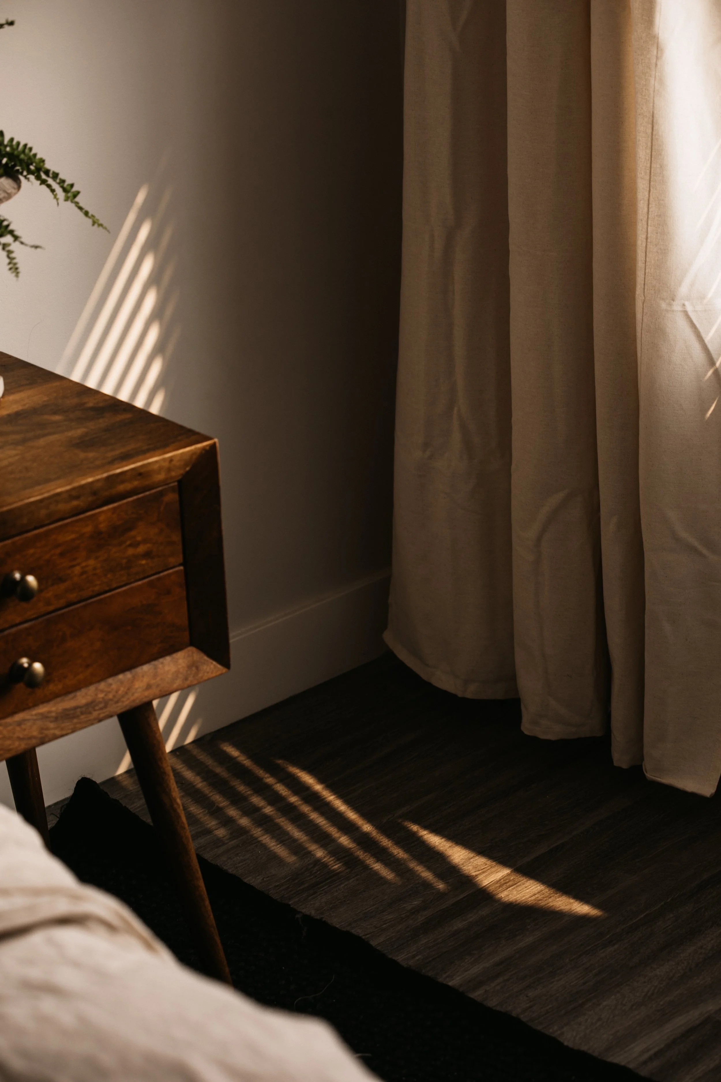 Sunlight streams through window blinds onto a beige curtain and dark wood floor in a cozy room, with part of a wooden bedside table visible.