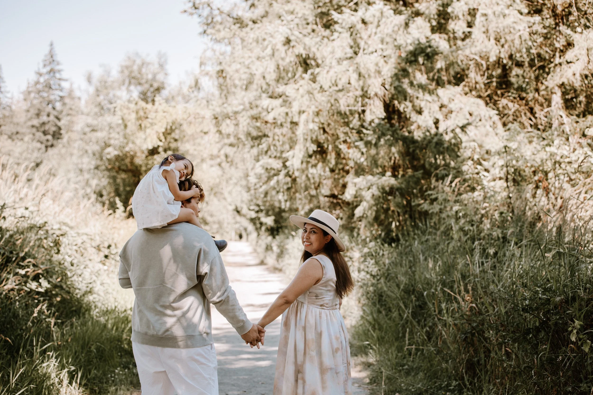 A family walking hand-in-hand on a dirt path surrounded by trees and greenery. The woman is wearing a dress and a wide-brimmed hat, the man is in casual clothes, and a girl is sitting on the man's shoulders, smiling and hugging him.