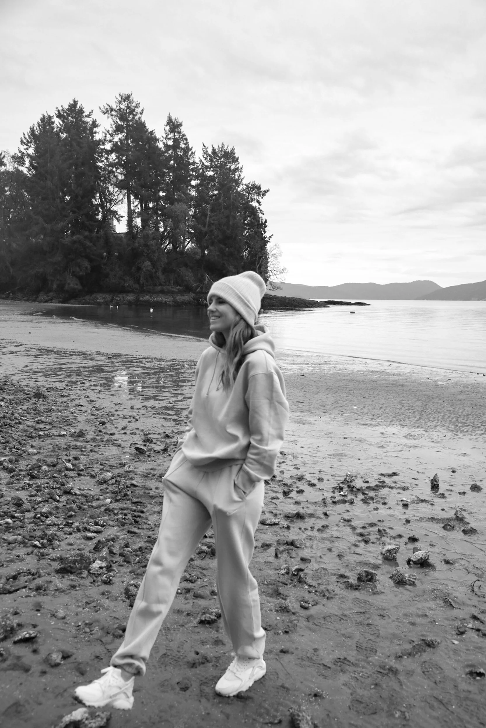 A woman in a hoodie and sweatpants wearing a beanie stands on a beach with trees and water in the background, captured in black and white.