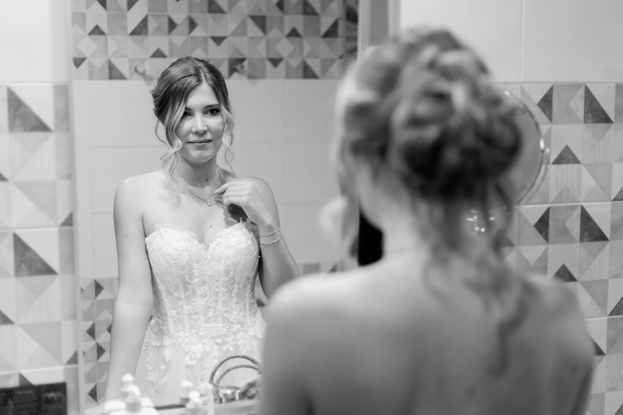 A bride looking at herself in a mirror, adjusting her necklace, with her back to the camera, in a bathroom with patterned tiles.