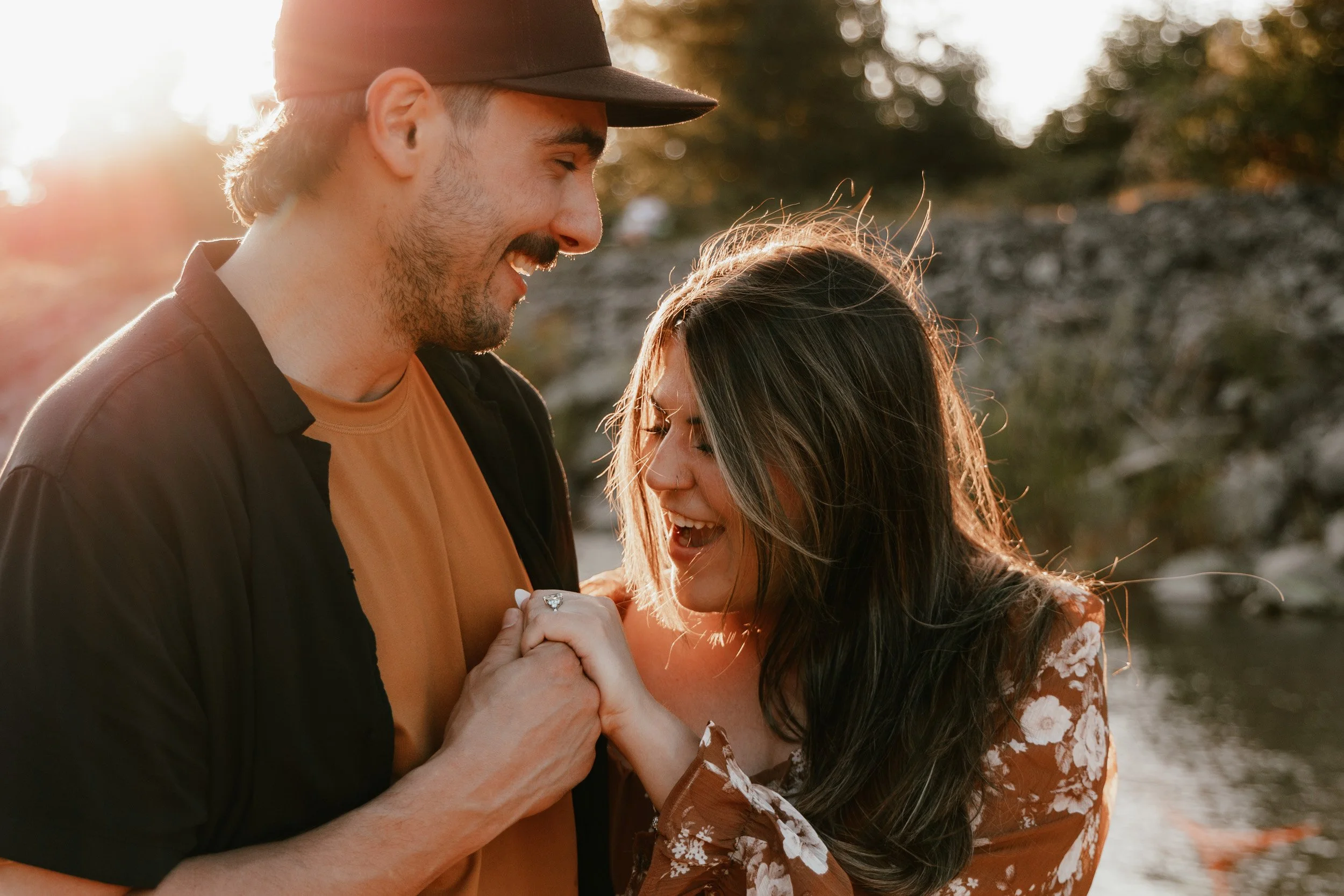 A smiling man and woman holding hands outdoors during sunset, with the sun shining in the background and trees behind them.